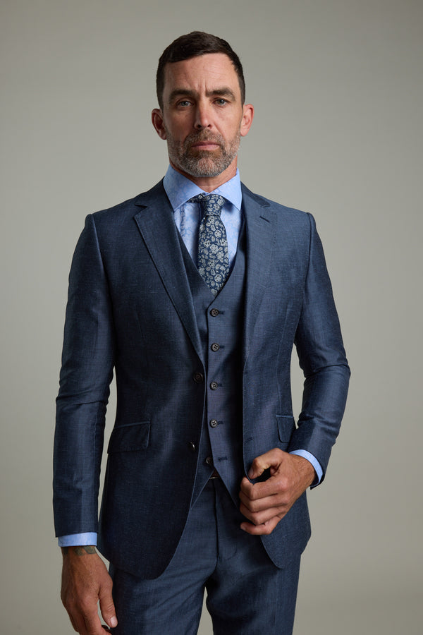 A man with short dark hair and a trimmed beard wears the Barkers Charles Texture Suit Jacket, a blue linen-wool three-piece, paired with a light blue dress shirt and patterned tie—ideal modern tailoring for summer weddings.