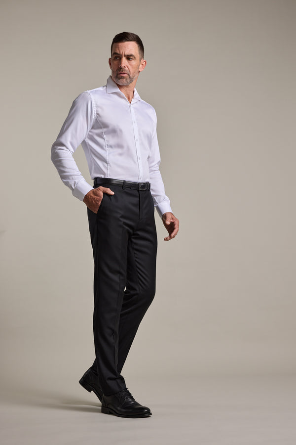A man stands against a plain background, wearing a white button-up shirt, Barkers' Mayfair ZQrx Suit Trouser in black crafted from ethical merino, and black shoes. One hand is in his pocket and he wears a serious expression.