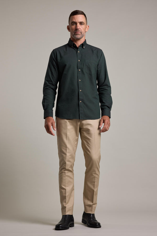 A man stands facing forward, wearing Lyndon Cotton Pants by Barkers in beige, a dark green button-up shirt, and black shoes, against a plain light gray background.
