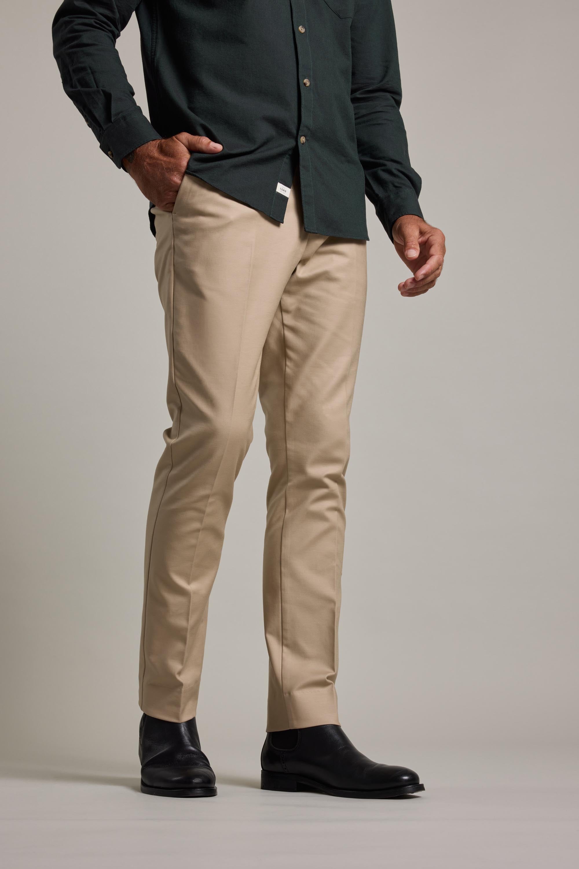 Wearing Barkers Lyndon Cotton Pants in beige, a dark green button-up shirt, and black dress boots, a person stands with one hand in their pocket against a plain background, exemplifying a smart casual style.