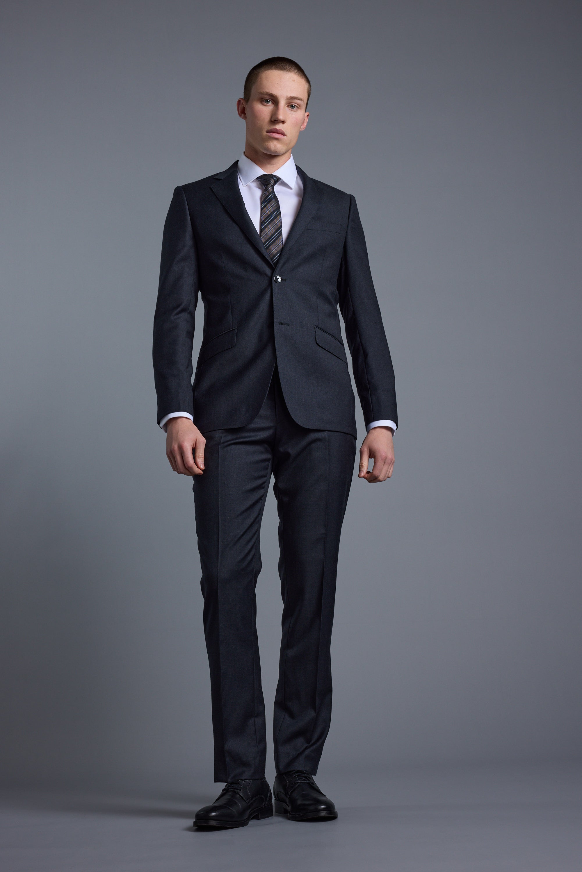 A man with short hair and a neutral expression stands against a gray background wearing the Barkers Mayfair ZQ Suit Jacket, crafted from breathable ZQrx Merino Wool. His arms are relaxed at his sides.