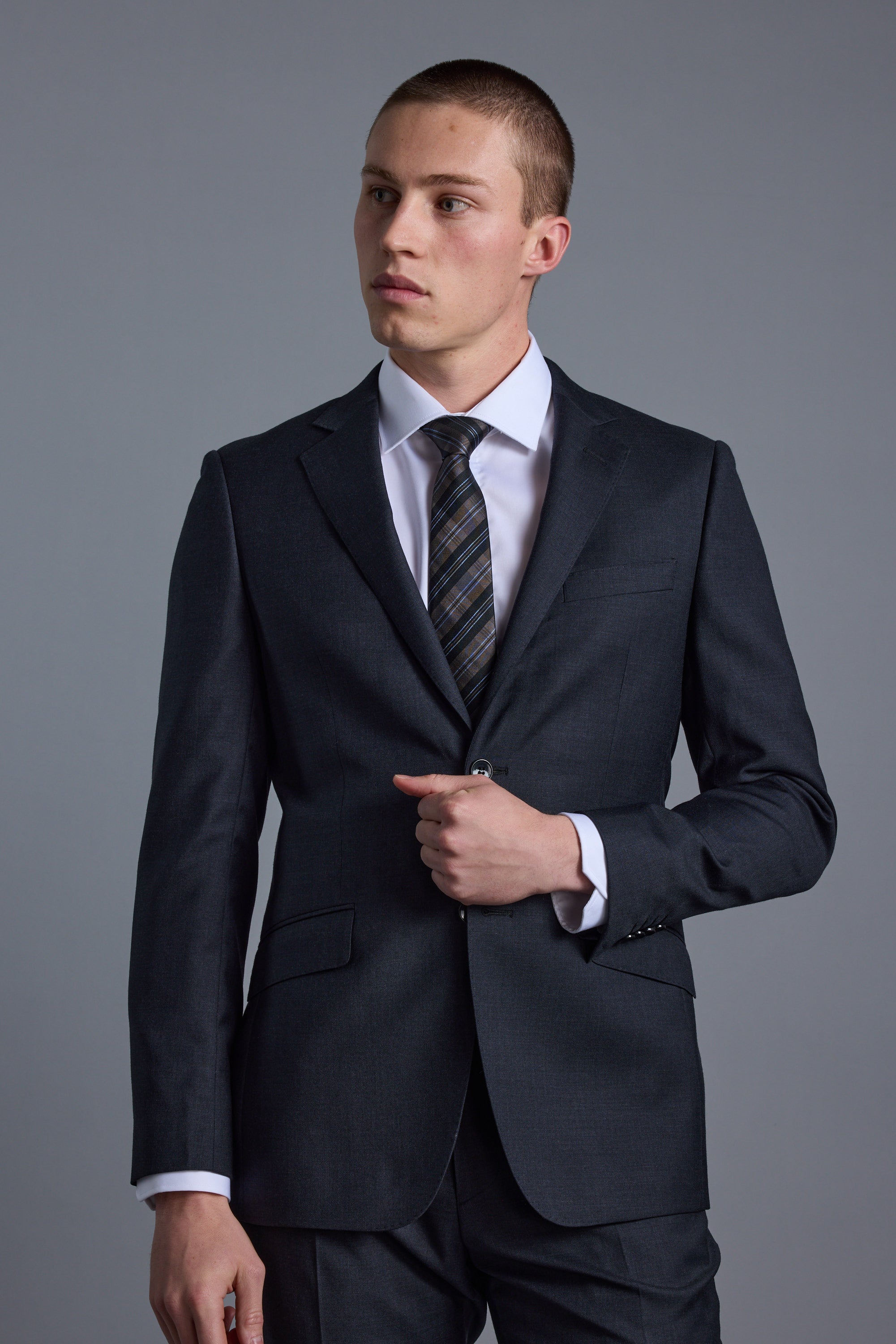 A young man in a dark Barkers Mayfair ZQ Suit Jacket stands against a plain gray background, wearing a white dress shirt and striped tie, looking slightly left with one hand adjusting his jacket.