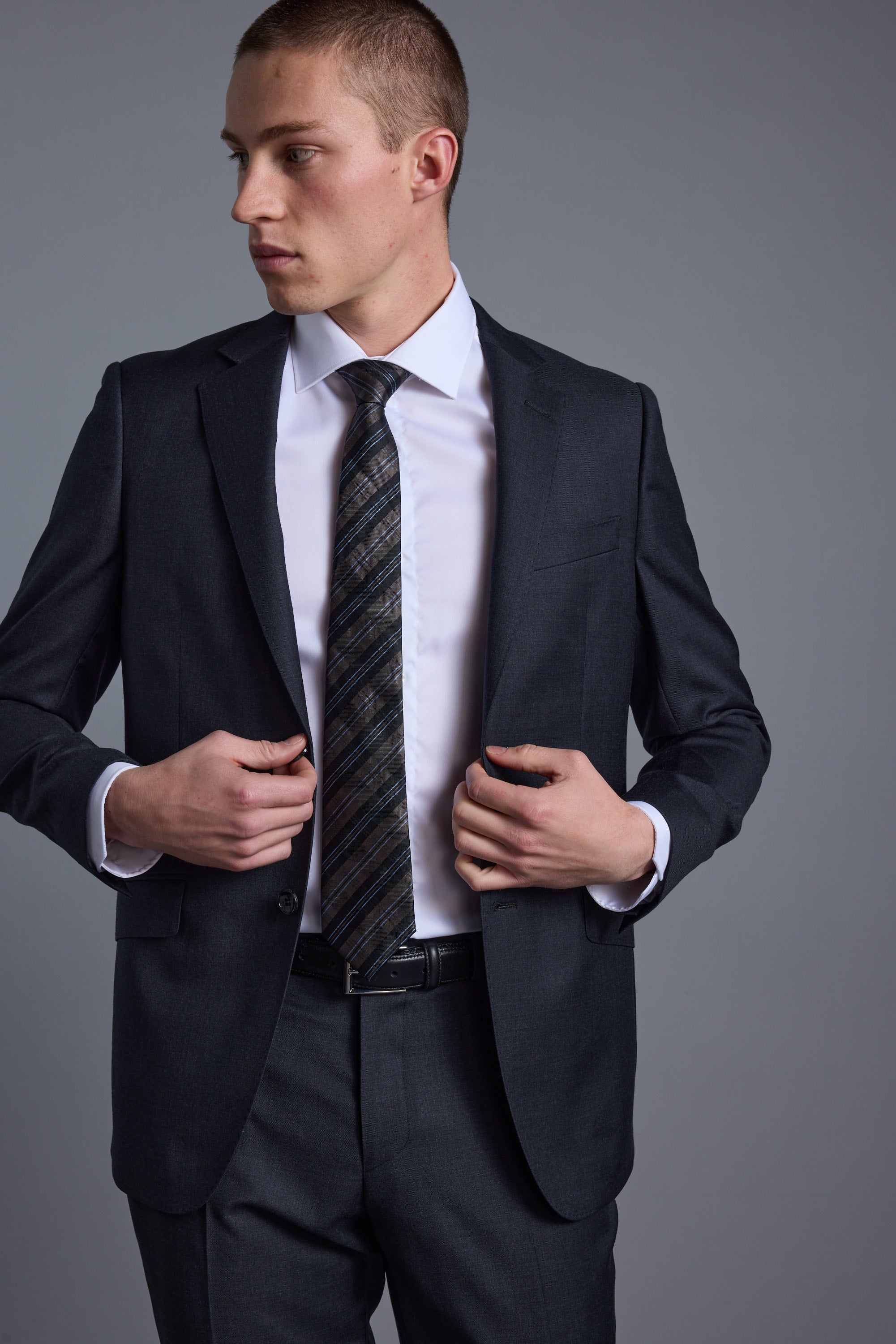 A man wearing the Barkers Mayfair ZQ Suit Jacket, crafted from breathable wool, stands against a plain gray background, looking to his left and adjusting his jacket with both hands.