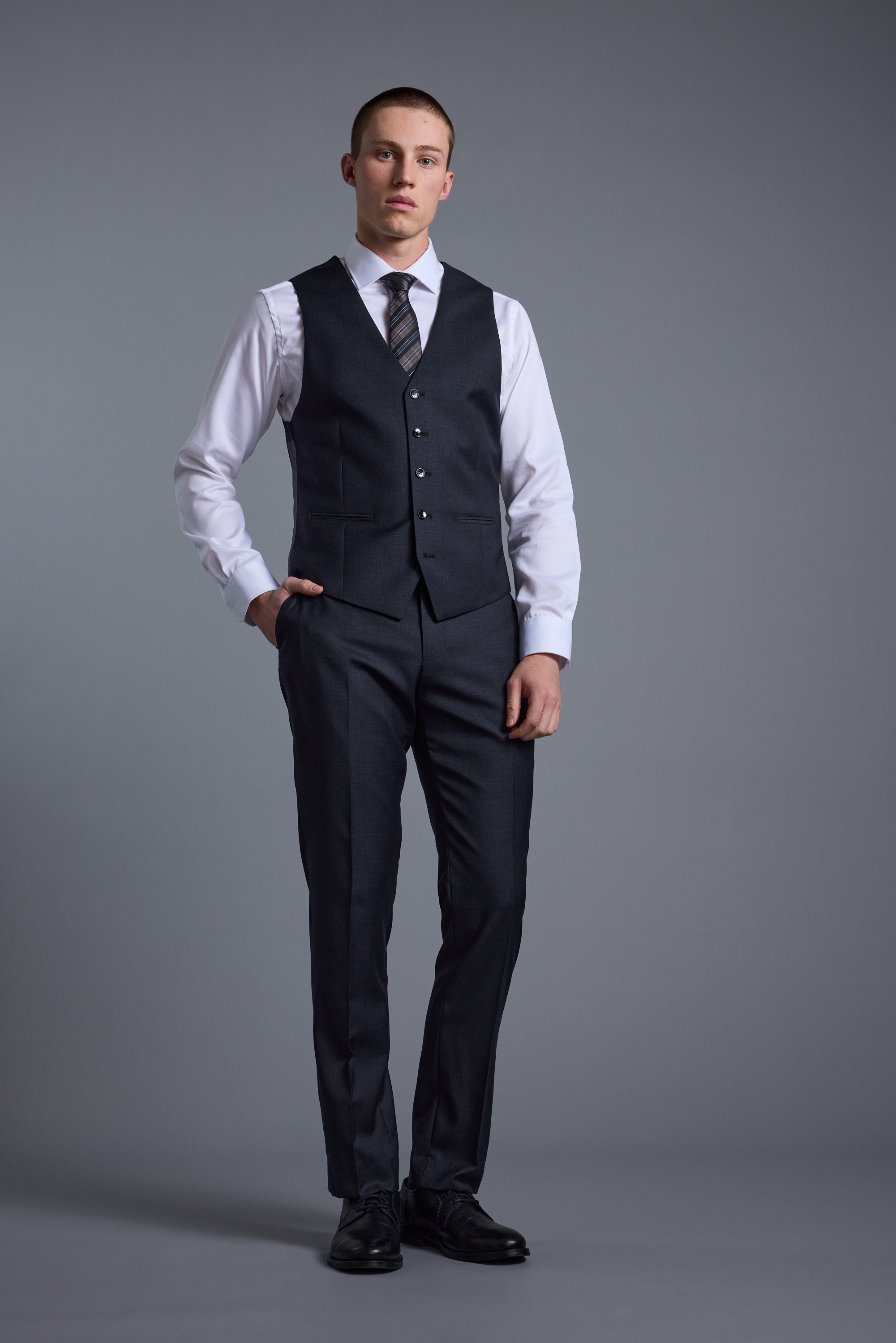 A man stands against a gray background wearing Barkers' Mayfair ZQ Suit Trousers, paired with a matching dark jacket, white dress shirt, patterned tie, and black shoes; one hand is in his pocket and he has a neutral expression.