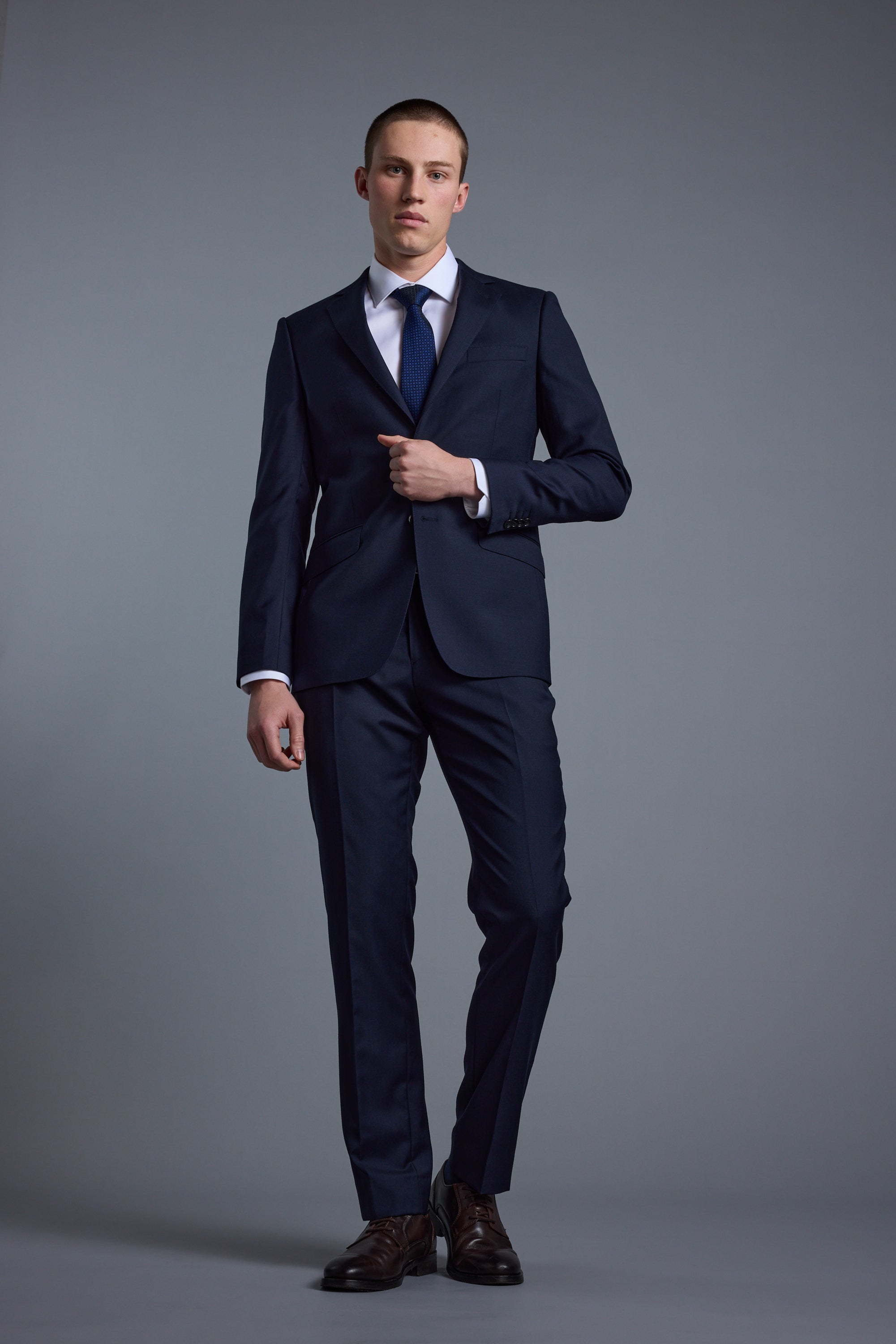 A man stands against a gray background wearing the Barkers Mayfair ZQ Suit Jacket with a white dress shirt, blue tie, and brown shoes. He faces forward, adjusting his suit jacket.