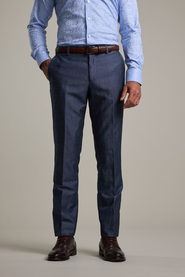 A man stands with one hand in his pocket against a plain background, wearing a light blue patterned shirt, Barkers Charles Texture Suit Trouser in dark blue, a brown belt, and brown dress shoes.