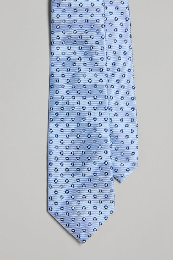 The Barkers Manhattan Flower Tie features light blue silk with a small dark blue and white circular pattern. Shown on a plain light gray background, it’s ideal for pairing with pocket squares.