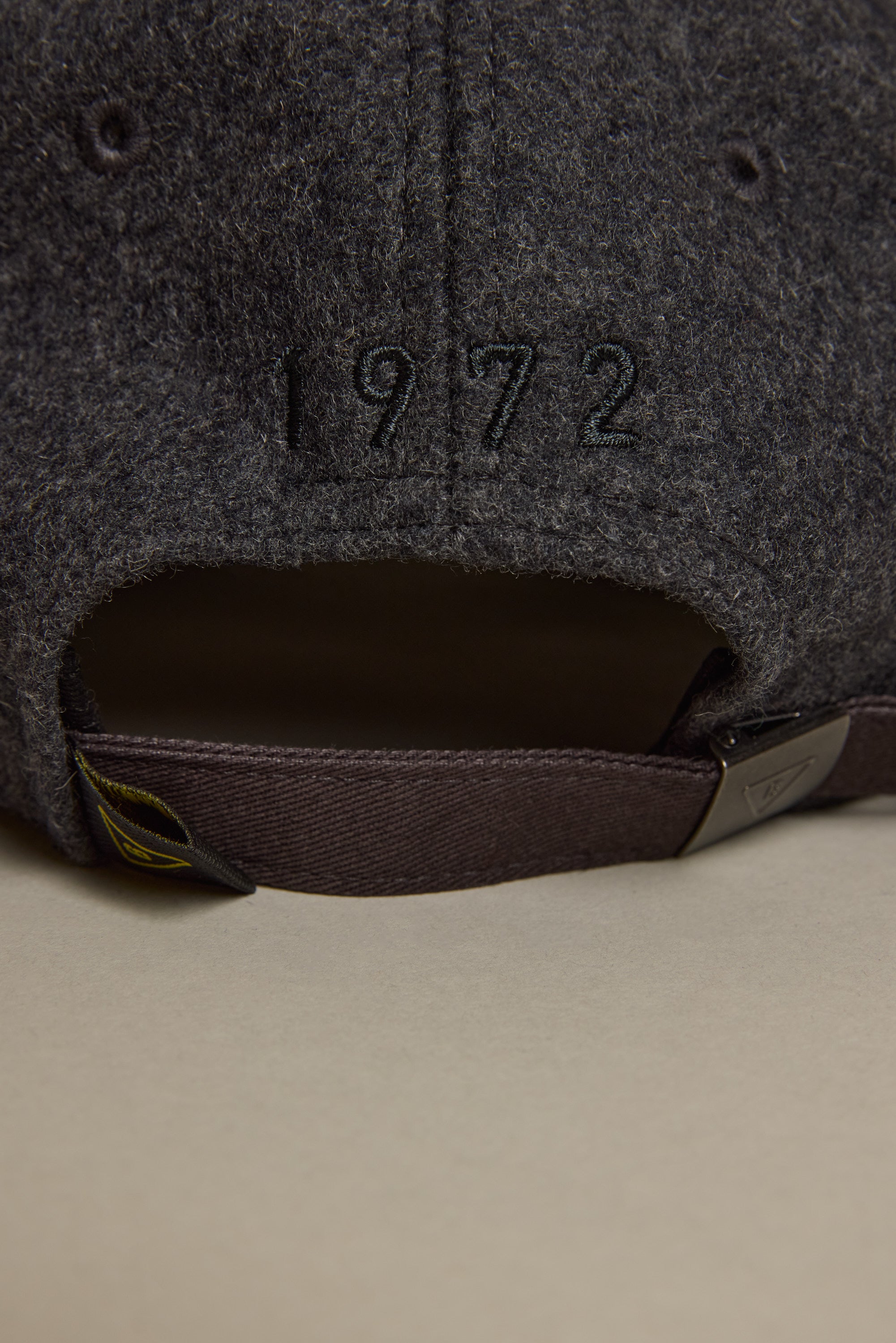 Close-up of the back of the Barkers Darcy Woollen Cap in dark gray wool, featuring "1972" embroidery above a brown adjustable strap with metal clasp. Cap rests on a light beige surface.