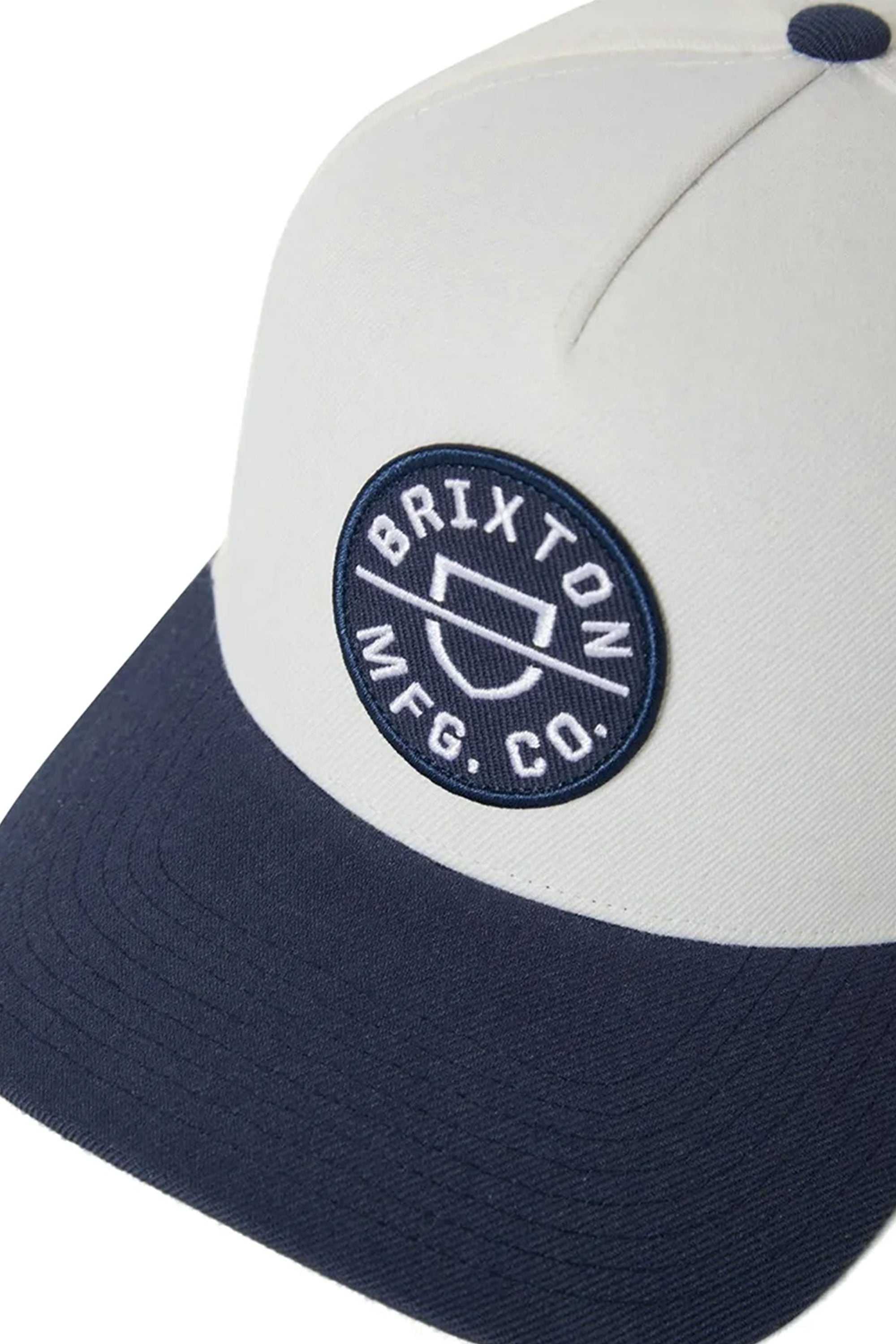 A close-up of the Brixton Crest Snapback by Brixton, a two-tone cap with a cream crown, navy brim, embroidered patch logo on the front, and a medium profile fit.