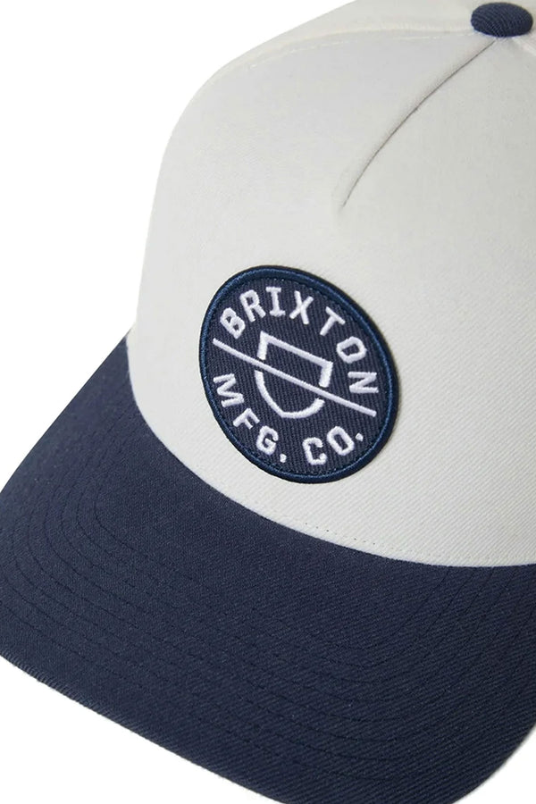 A close-up of the Brixton Crest Snapback by Brixton, a two-tone cap with a cream crown, navy brim, embroidered patch logo on the front, and a medium profile fit.