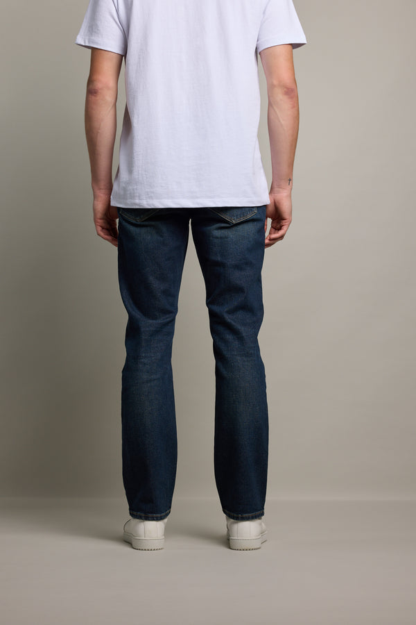 A person stands facing away from the camera, wearing a white t-shirt, Barkers Wanderer Denim Straight Jean made from sustainable denim, and white shoes, against a plain light background.