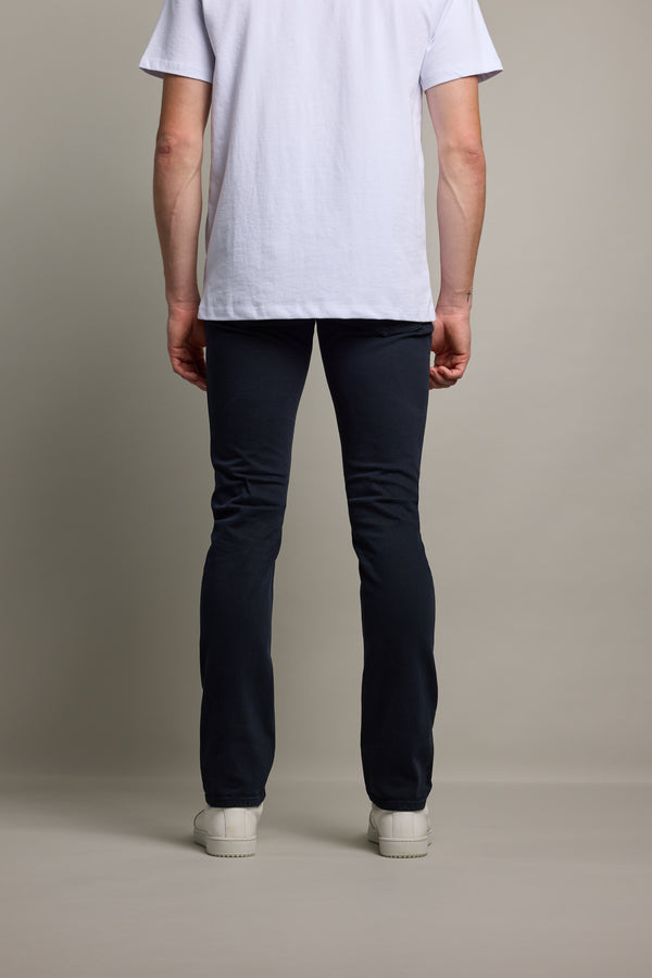 A person stands facing away from the camera, wearing a white T-shirt, Barkers Rambler Slim Coloured Jean made with regenerative cotton by Saitex Vietnam, and white sneakers, against a plain light background.