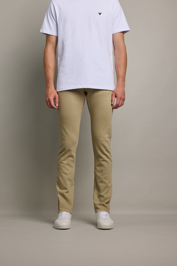 A person stands against a plain background in a white T-shirt, beige pants, and white sneakers. For a modern, eco-conscious look, pair with Barkers' Rambler Slim Coloured Jean. The image is cropped at the shoulders.
