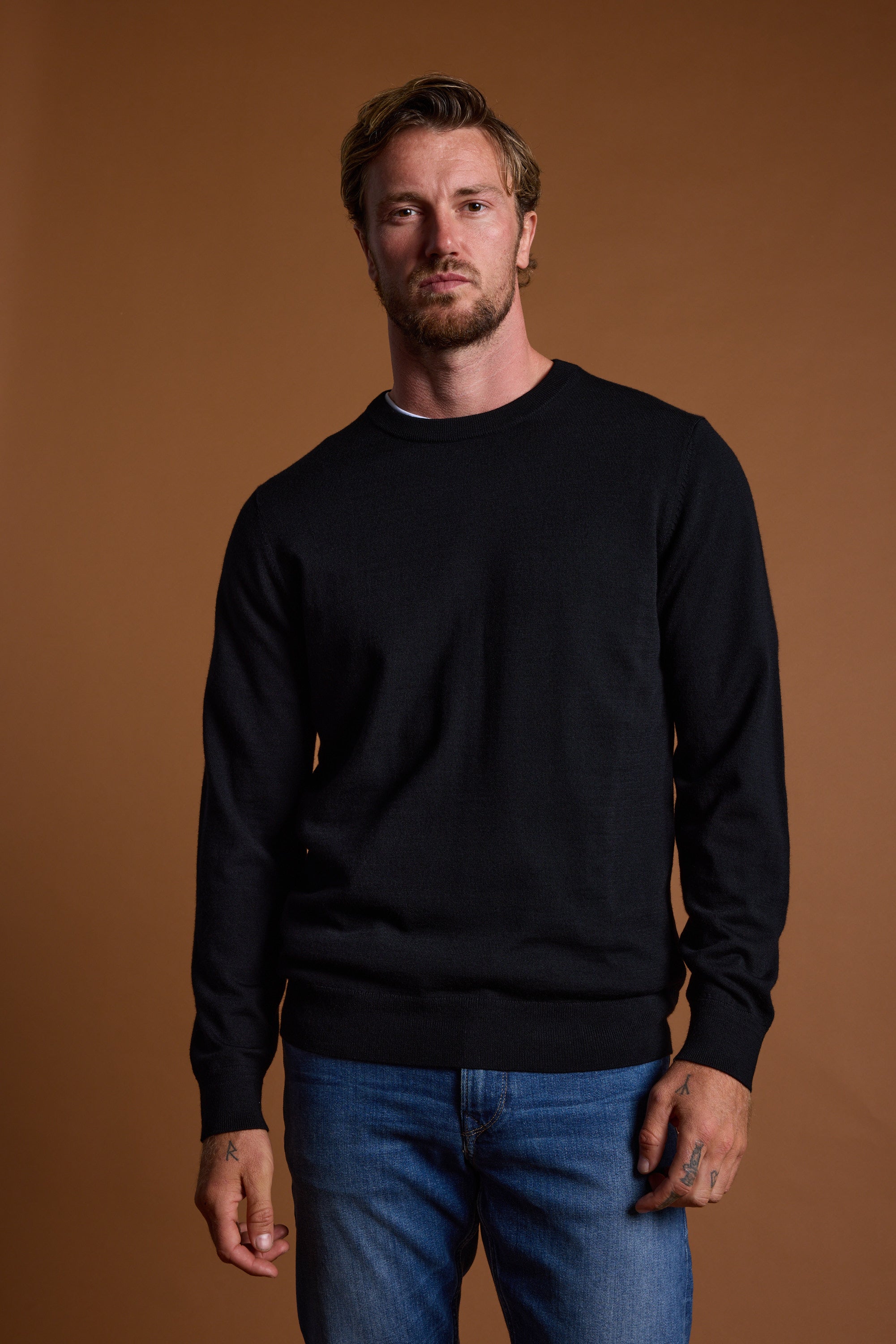 A man with short, light brown hair and a beard wears the Barkers Skyland ZQrx Merino Crew in black with blue jeans, standing against a plain brown background and showing a neutral expression.