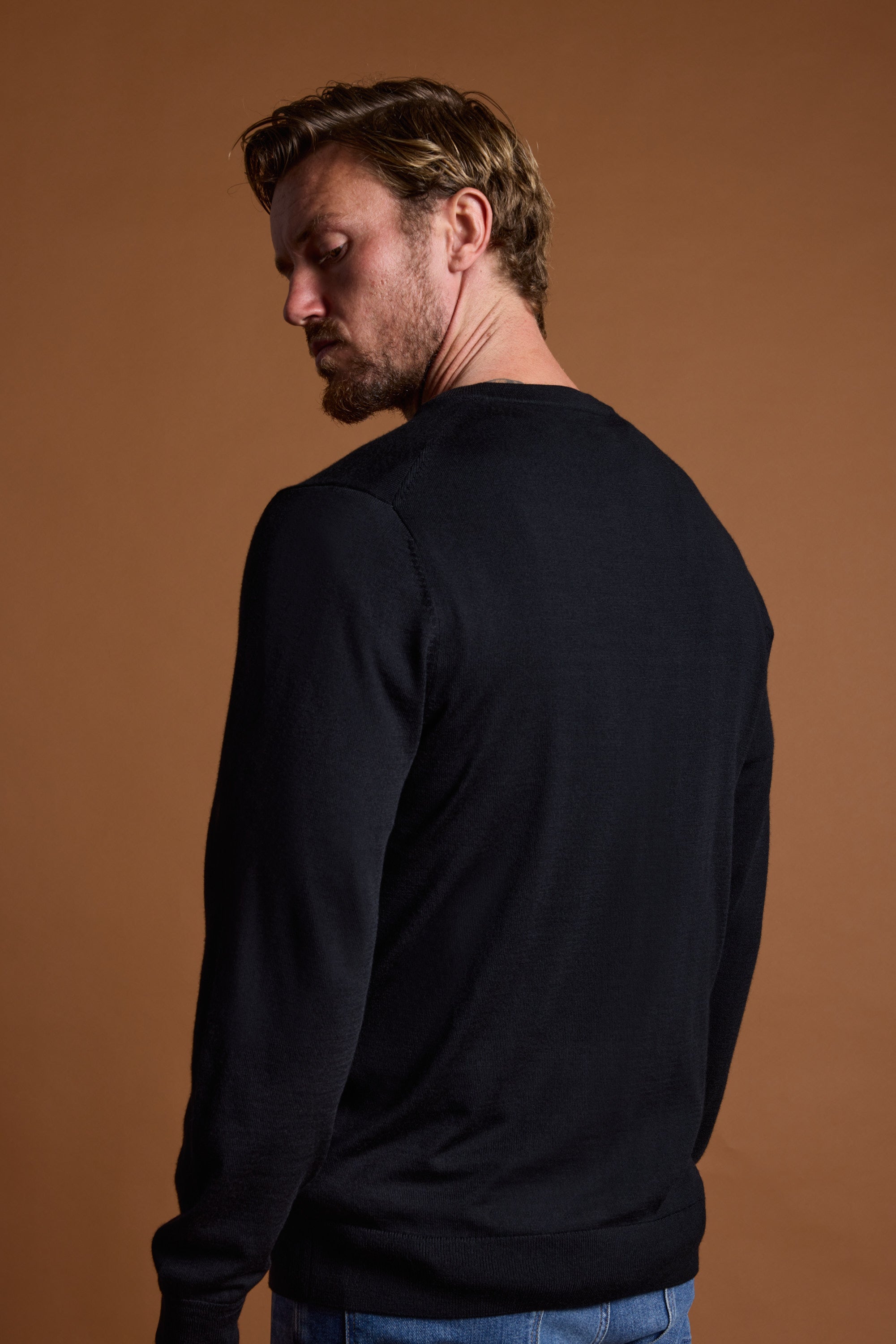 A man with light brown hair and a beard is wearing the Barkers Skyland ZQrx Merino Crew in black and blue jeans, standing against a brown background and looking over his left shoulder.