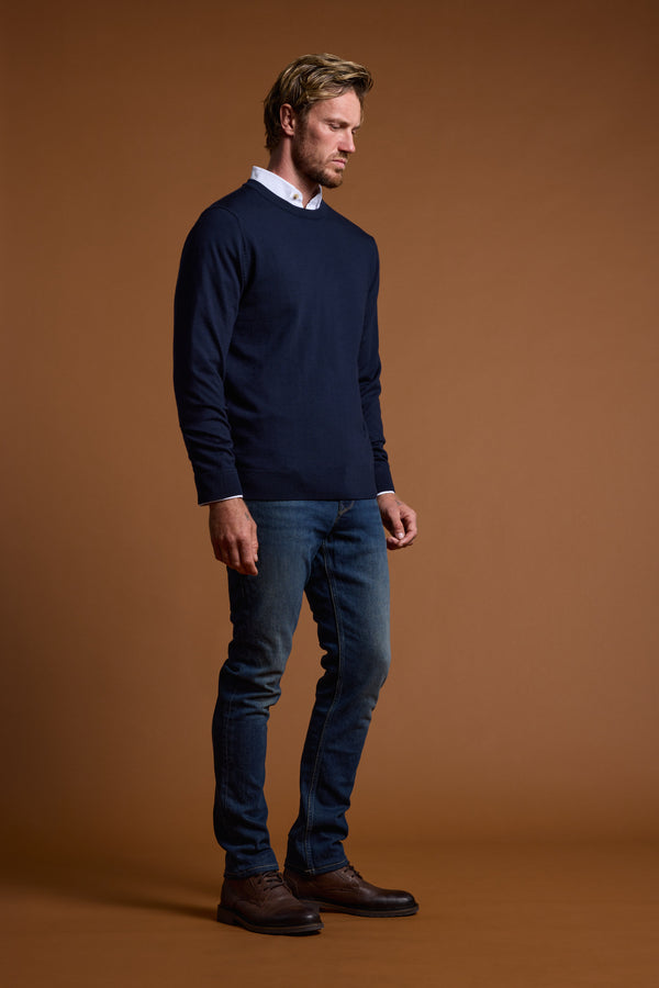 A man with short light brown hair and a beard stands against a brown background, wearing the Barkers Skyland ZQrx Merino Crew sweater over a white shirt, blue jeans, and brown shoes. He looks downward with a neutral expression.