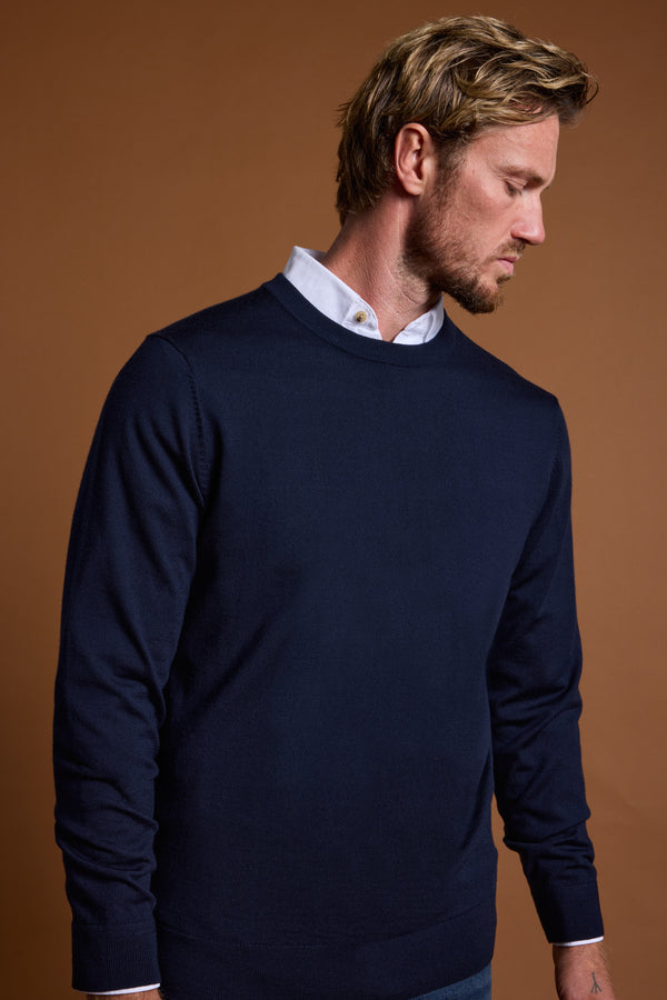 A man with short blond hair and a trimmed beard wears the Barkers Skyland ZQrx Merino Crew sweater in navy over a white collared shirt, standing against a plain brown background and looking down to his left.