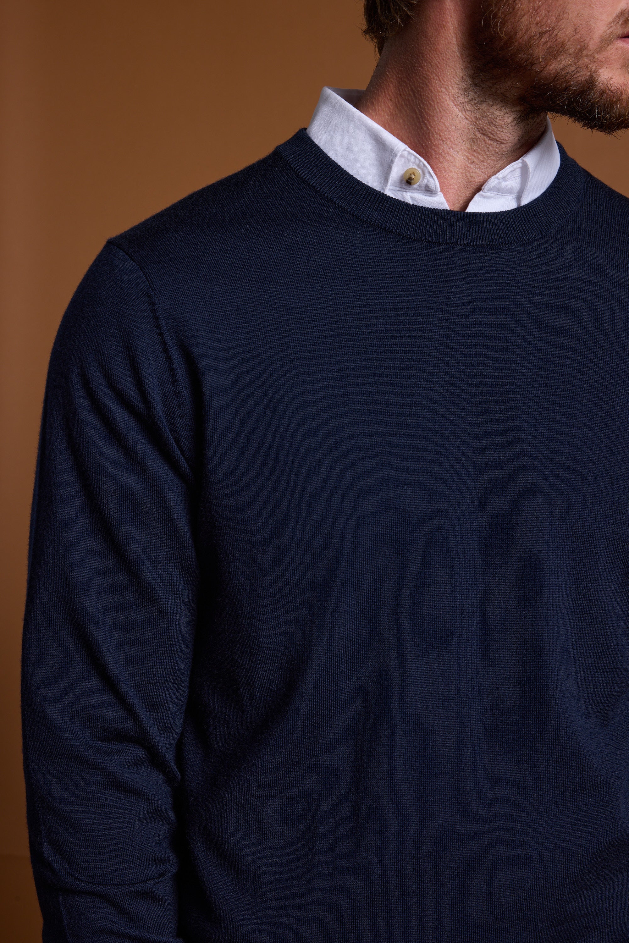 A man wearing the Barkers Skyland ZQrx Merino Crew in navy blue over a white collared shirt, shown from the shoulders down with part of his face out of frame, stands against a plain brown background.