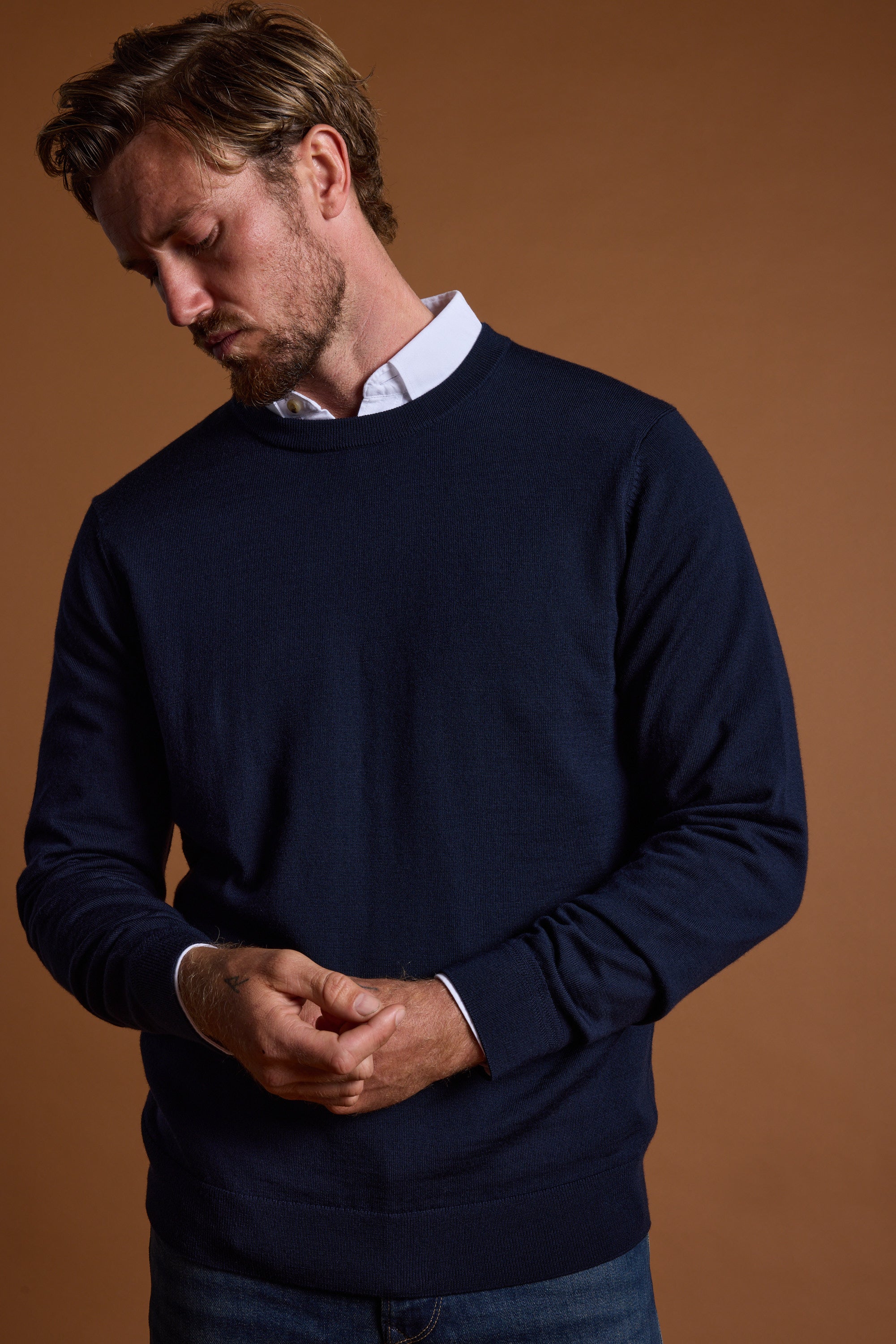 A man with light brown hair and a beard wears the Barkers Skyland ZQrx Merino Crew in navy over a white collared shirt and blue jeans, standing against a brown background with his hands loosely clasped, looking down.