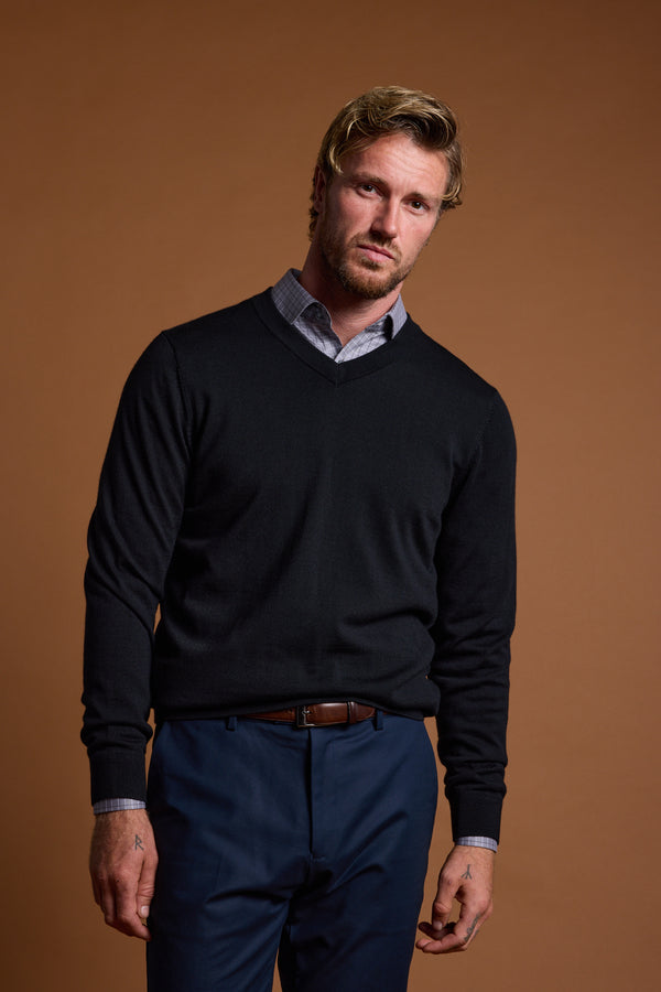 A man with blond hair and a short beard wears the Barkers Skyland ZQrx Merino Vee—a black, traceable merino V-neck sweater—over a collared shirt and blue pants. He stands against a plain brown background with a neutral expression and relaxed posture.
