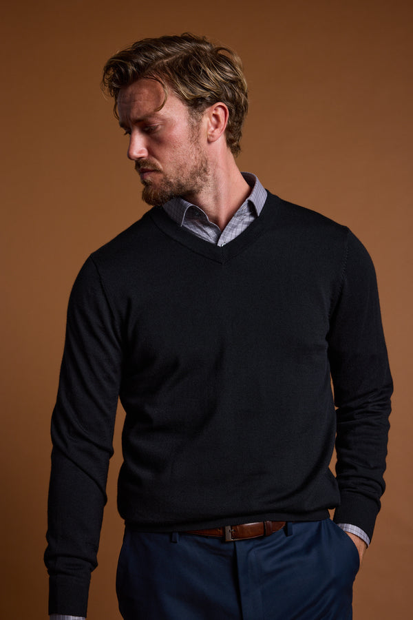A man with wavy hair and a beard looks left, wearing the Barkers Skyland ZQrx Merino Vee sweater over a collared shirt, paired with dark pants and a brown belt, standing against a plain brown background.