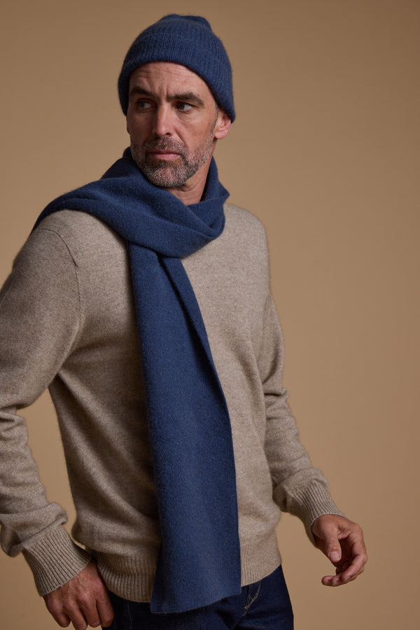 A man in a beige sweater accessorizes with the NZ Made Perino Beanie and matching scarf by Woolyarns Perino, both in a blue merino possum silk blend, standing against a tan background and looking to the side with a neutral expression.