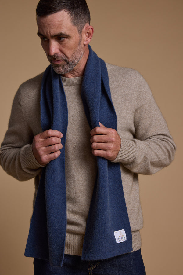 A man with short dark hair and a beard wears a beige sweater and holds the Woolyarns Perino NZ Made Perino Scarf in blue. The plain beige background highlights New Zealand’s sustainable fashion style.
