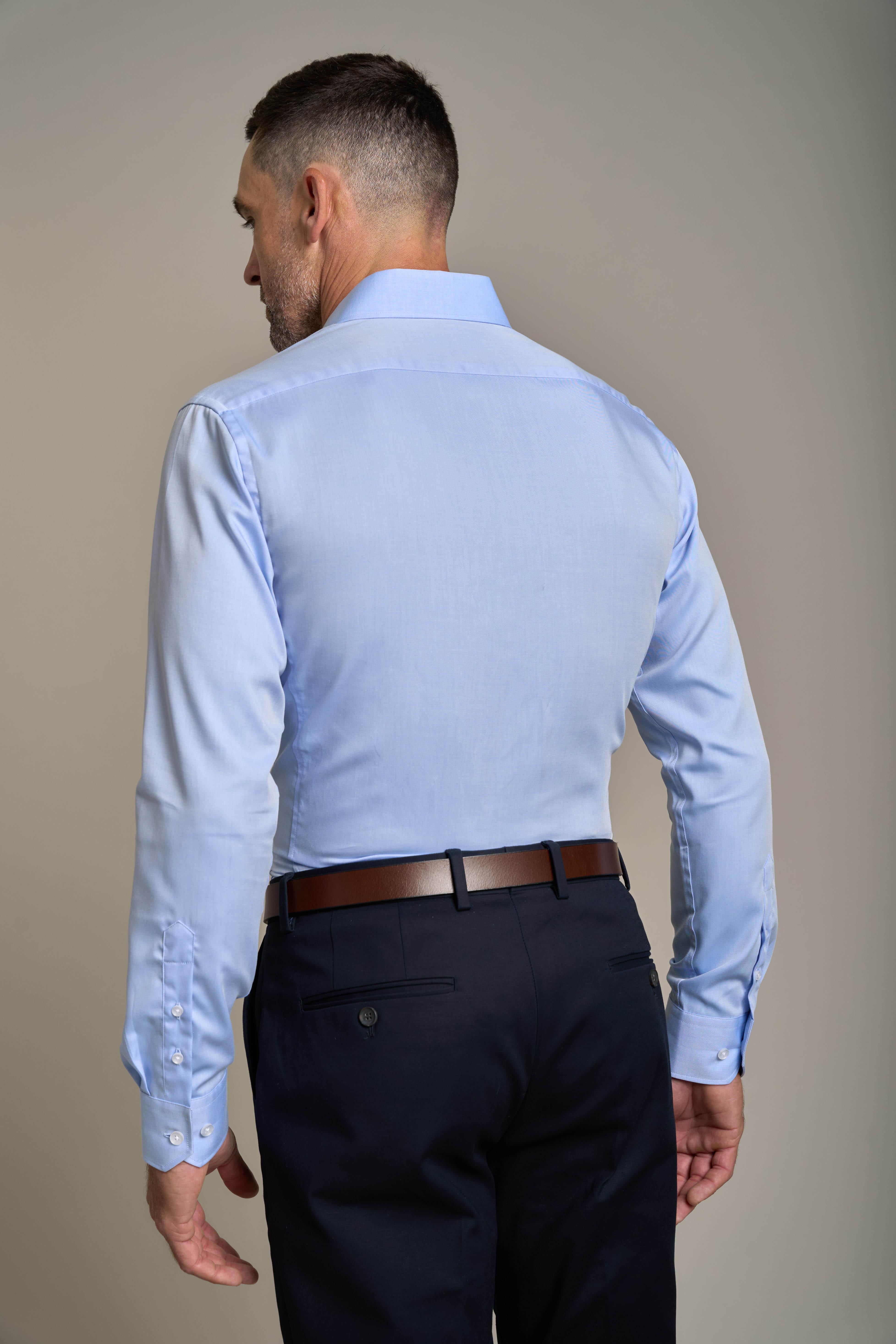 A man with short hair is shown from the back, wearing the Barkers Tribeca Reg Cuff Shirt in light blue, tucked into black dress pants with a brown belt, standing against a plain beige background.