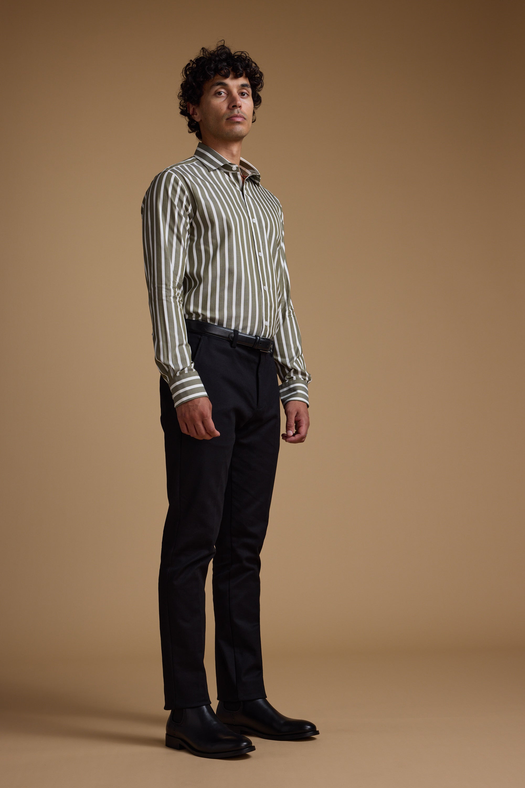 A man with curly hair stands against a plain brown background, wearing the Barkers Nandas Stripe Business Shirt in green and white, black trousers, and black shoes. He looks slightly upward with a neutral expression.