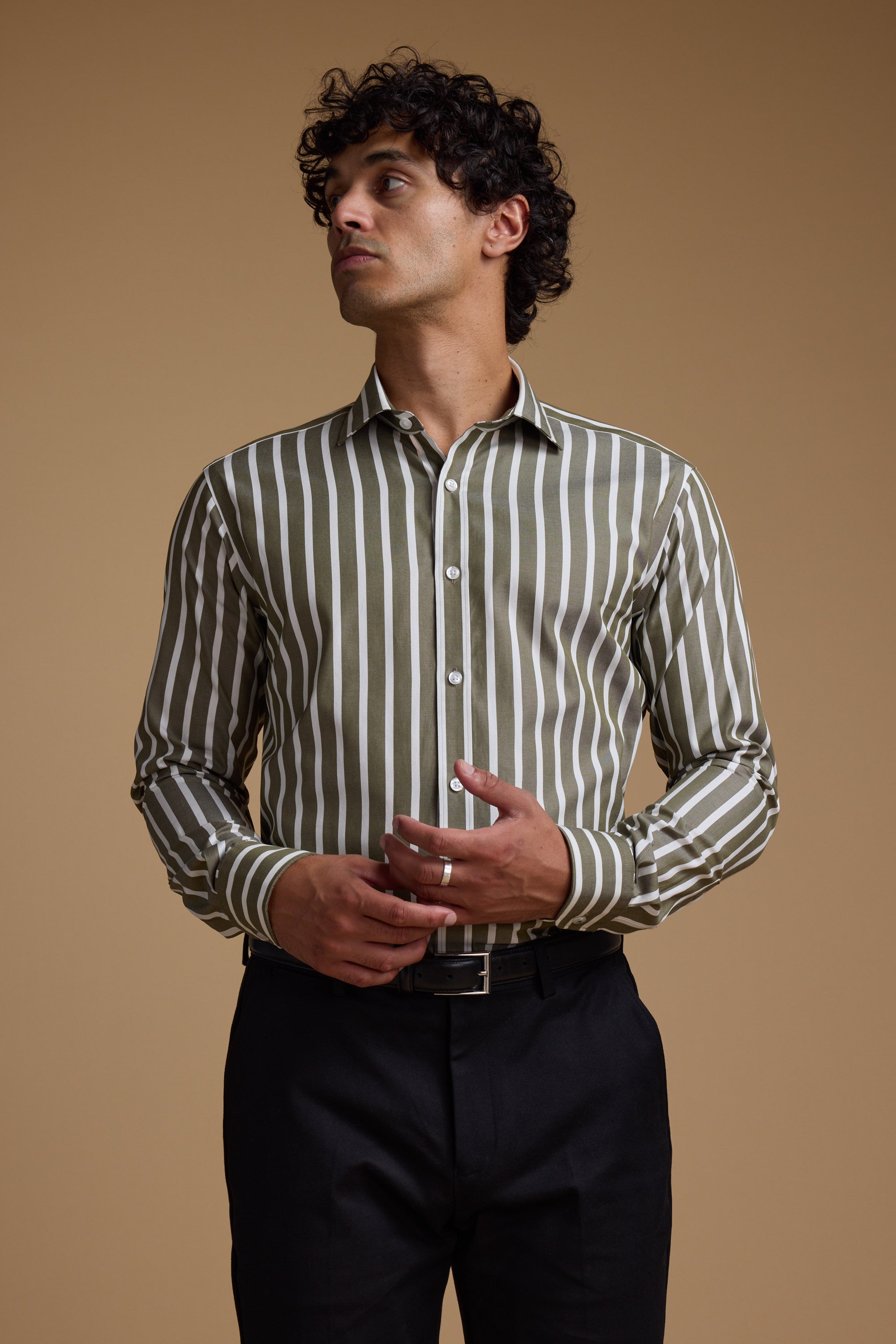 A man with curly hair wears the Barkers Nandas Stripe Business Shirt, featuring green and white vertical stripes, along with black pants. He stands against a plain beige background, looking to his left.