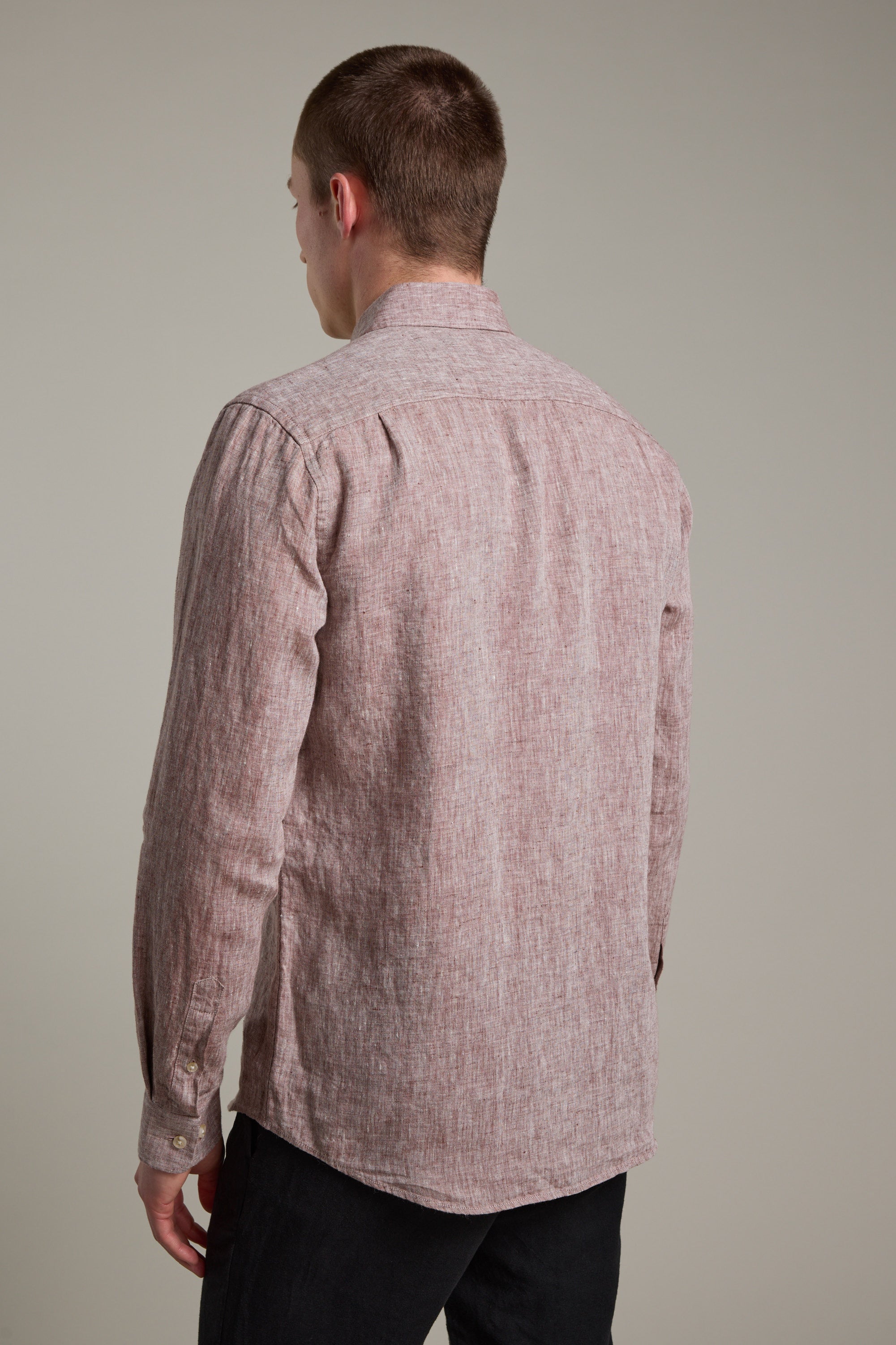 A person stands facing away from the camera, wearing Barkers' Dorset Linen Shirt—a long-sleeved, light red tailored-fit French linen shirt—with dark pants, set against a plain beige background.