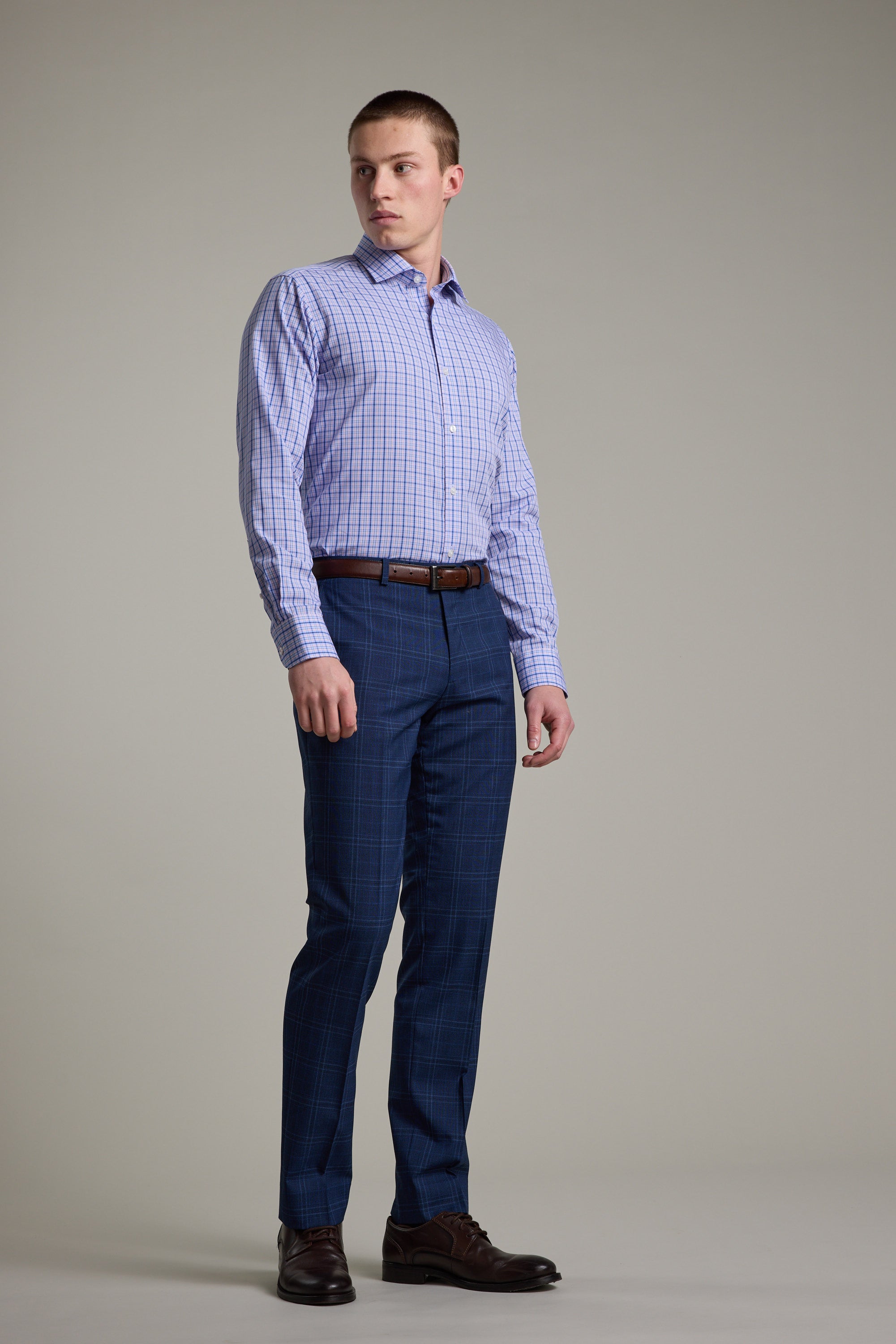 A man stands against a plain background wearing the Barkers Denton Check Business Shirt, blue plaid dress pants, a brown belt, and brown dress shoes. He looks slightly left with a neutral expression. The tailored cotton shirt completes his polished look.