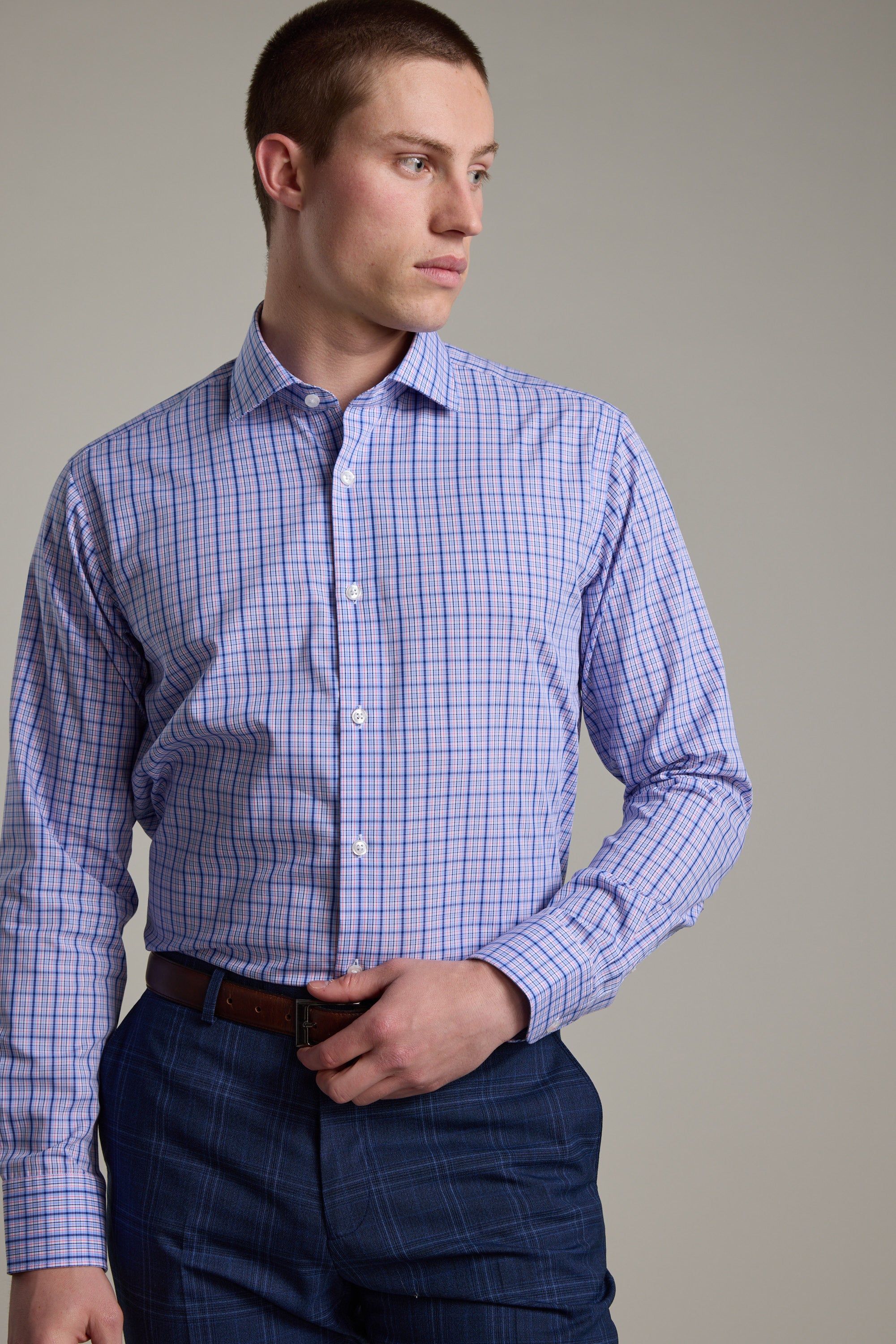 A man in a Barkers Denton Check Business Shirt, tailored fit in light purple and blue, paired with dark blue plaid pants and a brown belt, stands sideways with his left hand on his waist, gazing to the right.