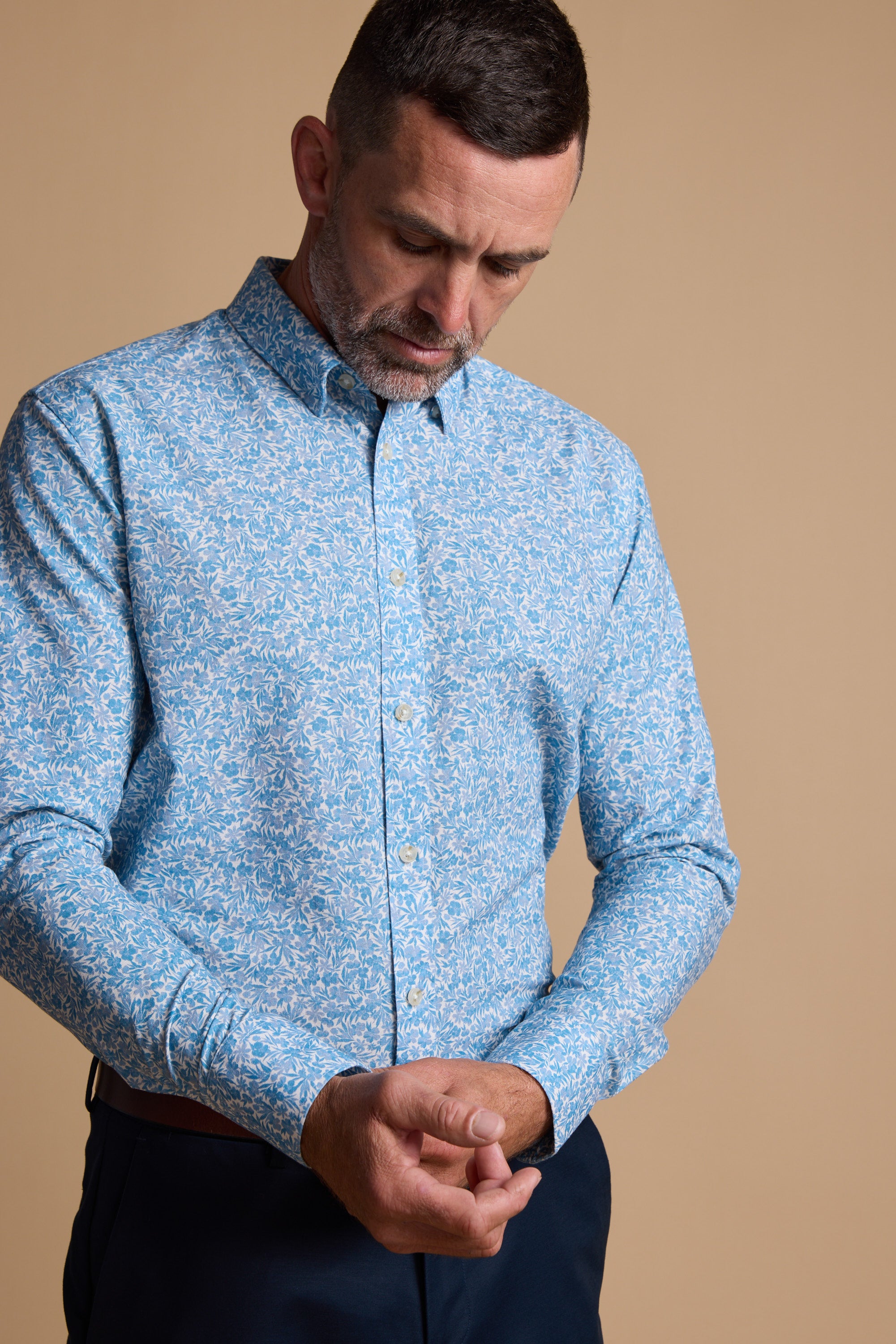 A man in the Barkers Torrey Print Shirt, a light blue patterned button-up crafted from responsibly sourced cotton, fastens his sleeve while looking down against a plain beige backdrop.