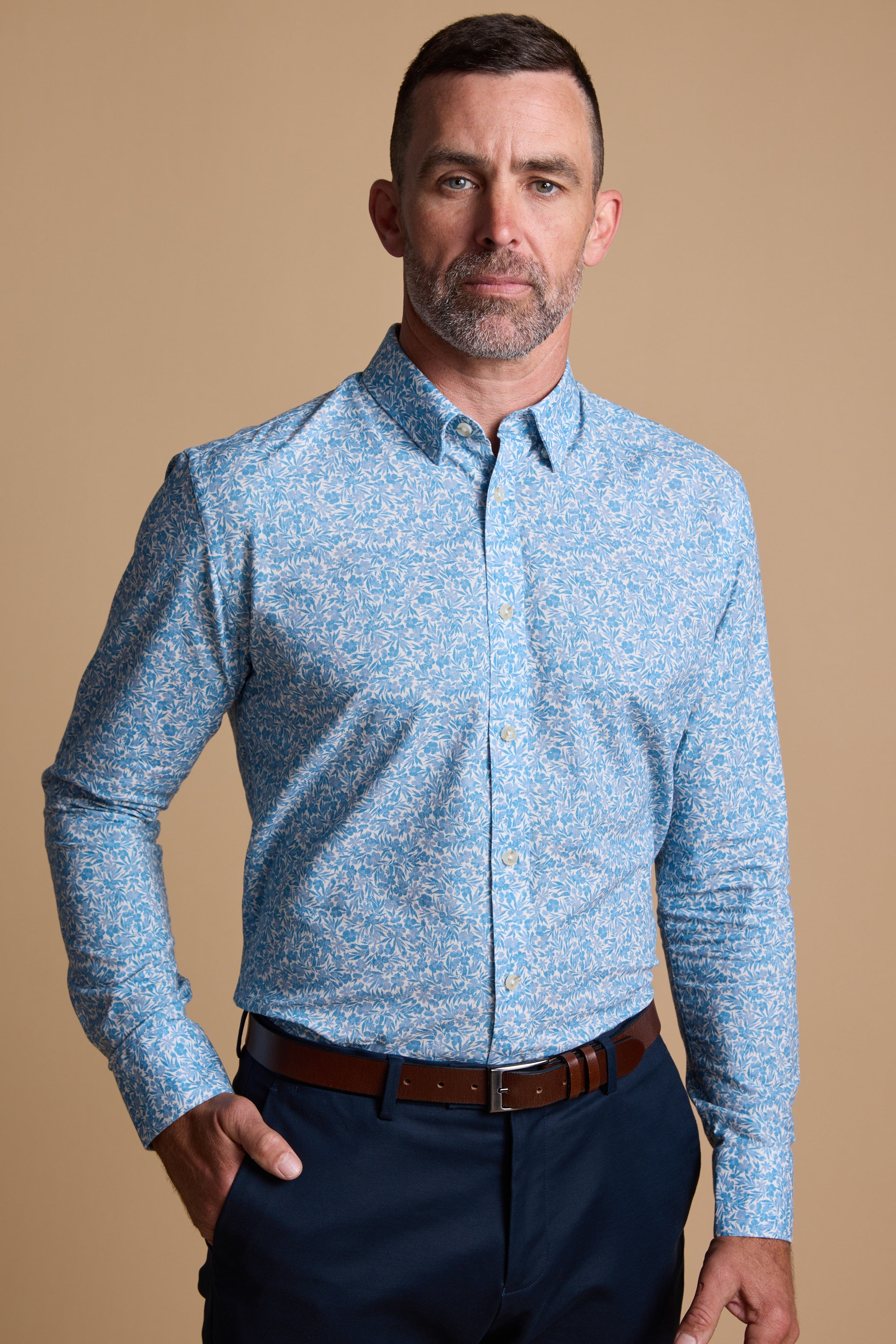 A man with short hair and a beard wears the Barkers Torrey Print Shirt in light blue—made from responsibly sourced cotton—with dark pants and a brown belt, standing against a plain beige background.