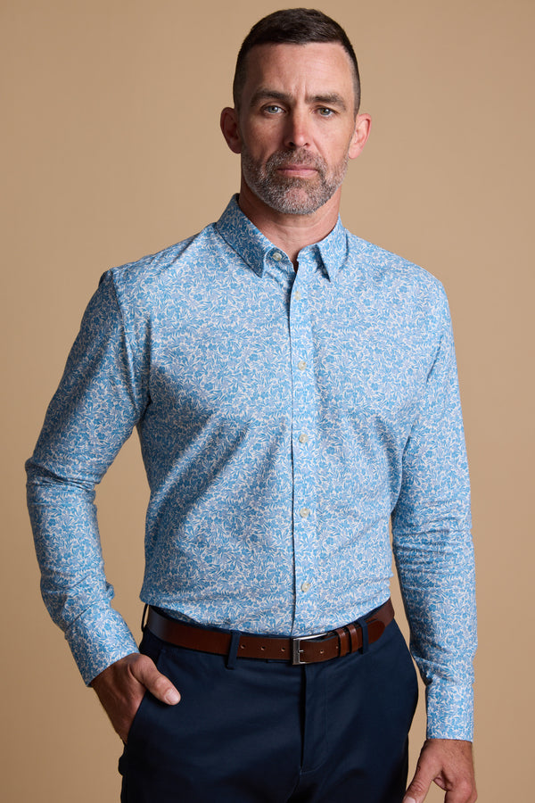 A man with short hair and a beard wears the Barkers Torrey Print Shirt in light blue—made from responsibly sourced cotton—with dark pants and a brown belt, standing against a plain beige background.