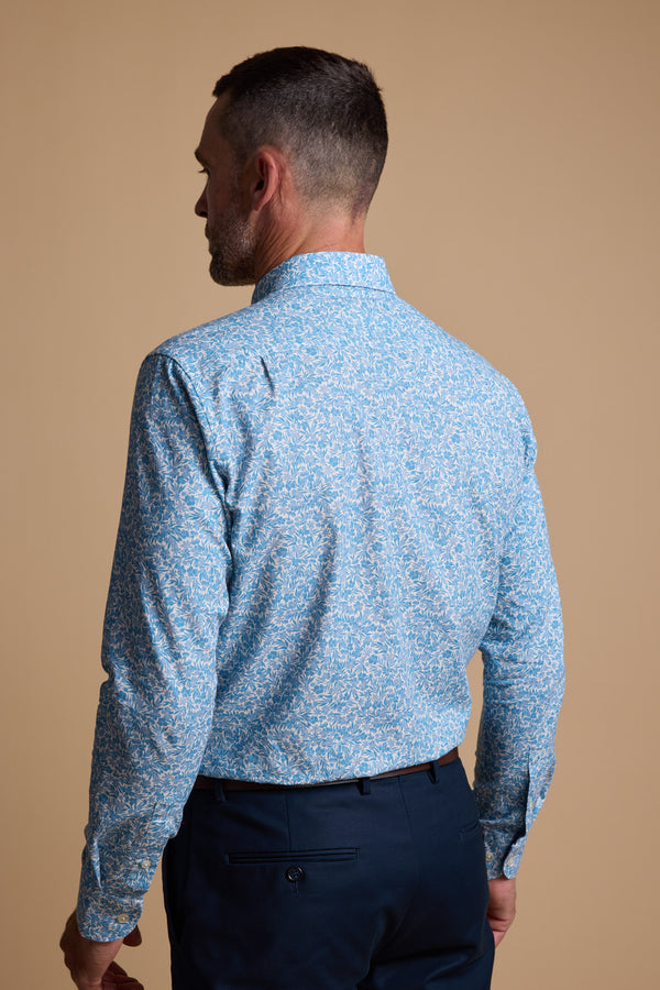 A man with his back to the camera wears a light blue Barkers Torrey Print Shirt featuring a subtle floral pattern, tucked into dark blue trousers, and stands against a beige background.