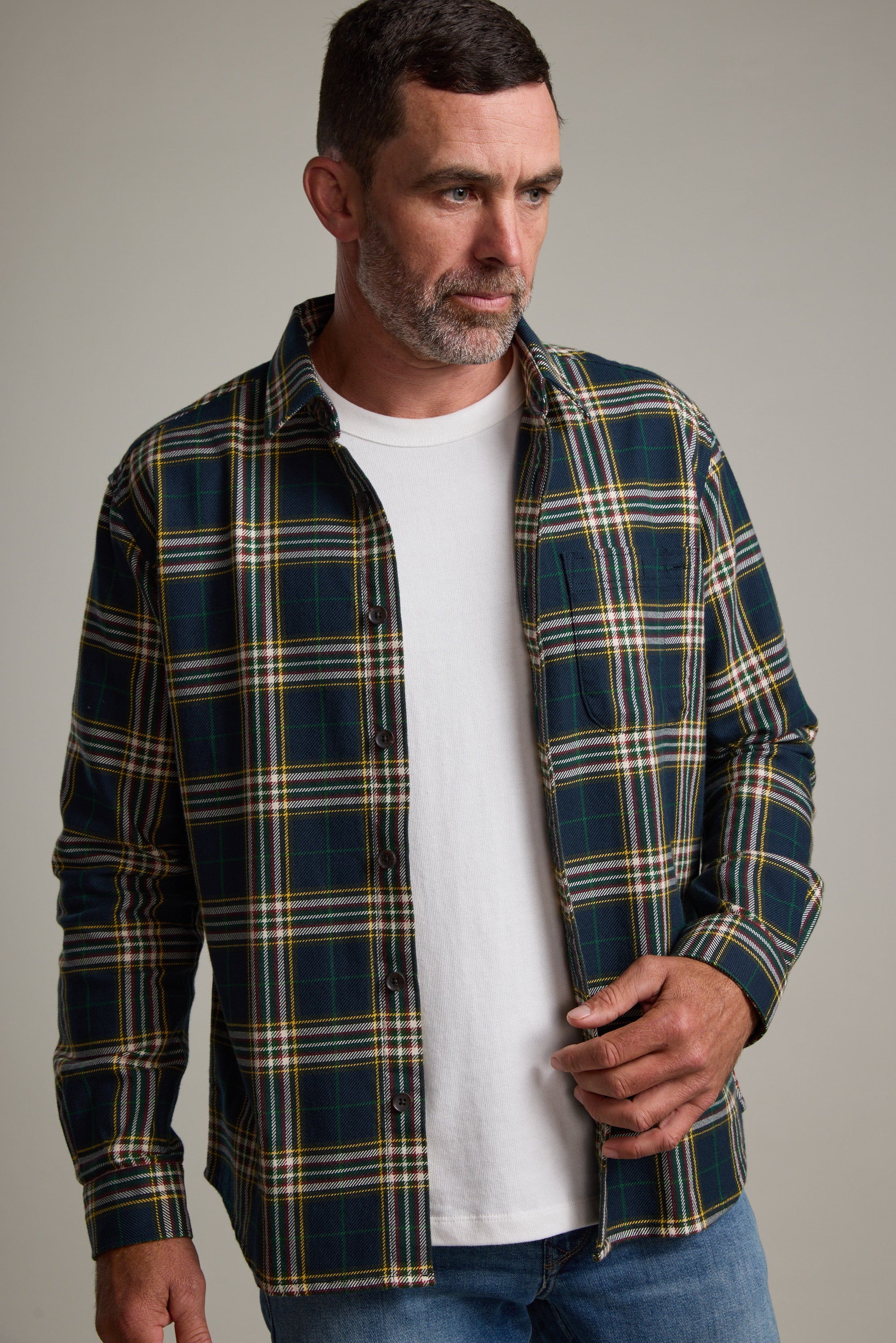 A man with short dark hair and a beard wears the Barkers Brennan Check Overshirt in green and yellow, made from responsibly sourced cotton, over a white t-shirt and blue jeans against a plain light background.