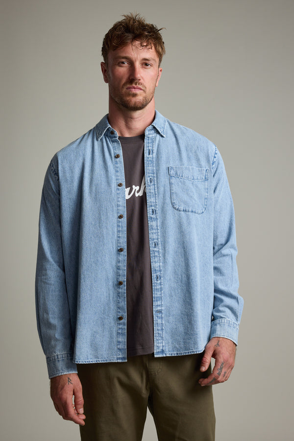 A man with short, tousled hair wears the Barkers Ruxton Denim Shirt as an overshirt over a black tee and olive green pants, standing against a plain gray background.
