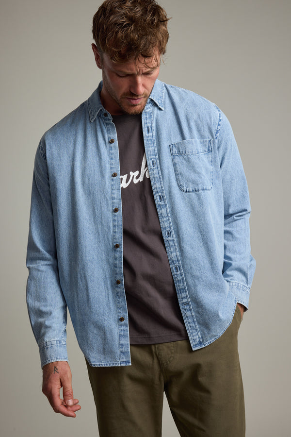 A man wears the Barkers Ruxton Denim Shirt in a classic fit, layered over a dark tee with white text and olive green pants, standing with head slightly bowed and one hand in his pocket against a neutral background.