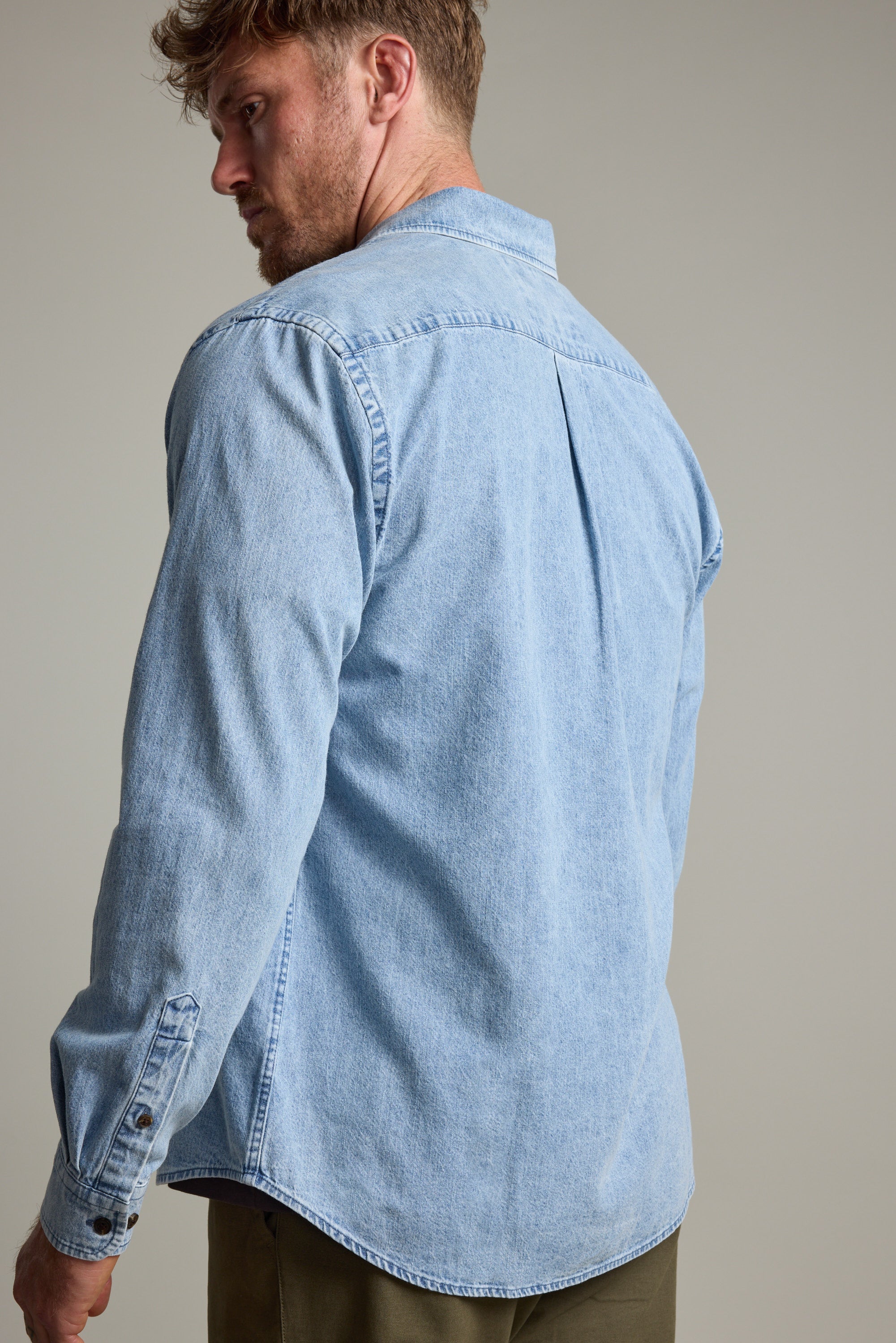 A man with light brown hair and a beard stands against a plain background, wearing the Barkers Ruxton Denim Shirt styled as an overshirt with olive green pants.