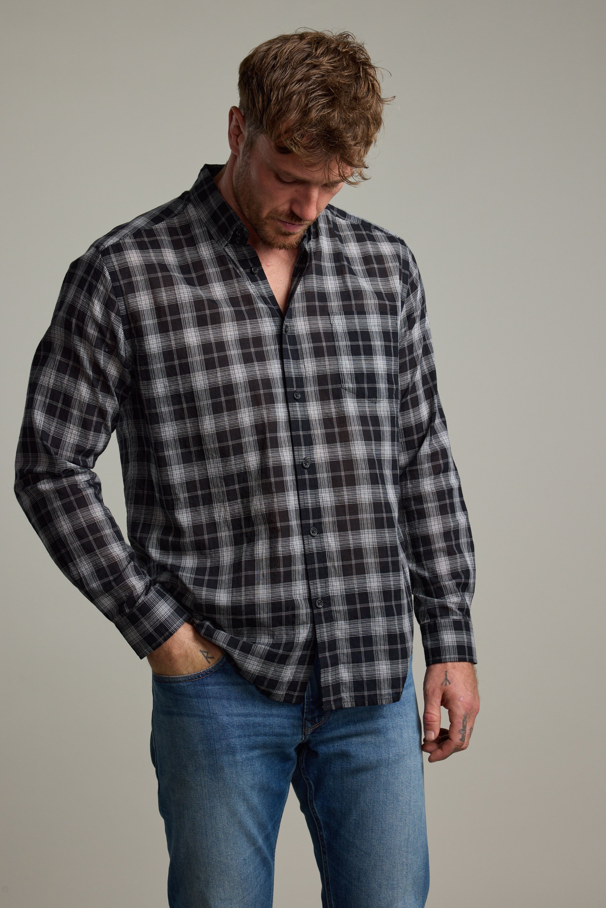 A man in the Barkers Carter Check Shirt, featuring a classic fit and black, white, and gray checks, pairs it with blue jeans and stands against a plain background, looking down with one hand in his pocket.