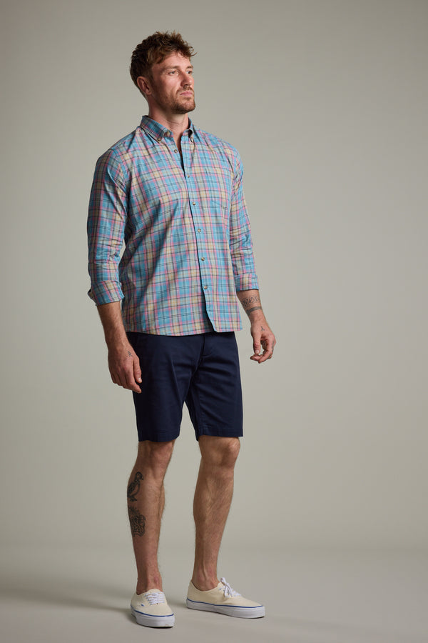 A man with short hair and visible tattoos stands against a plain background wearing the Barkers Hayne Check Shirt, rolled at the sleeves, navy blue shorts, and white slip-on shoes. He has a neutral expression.