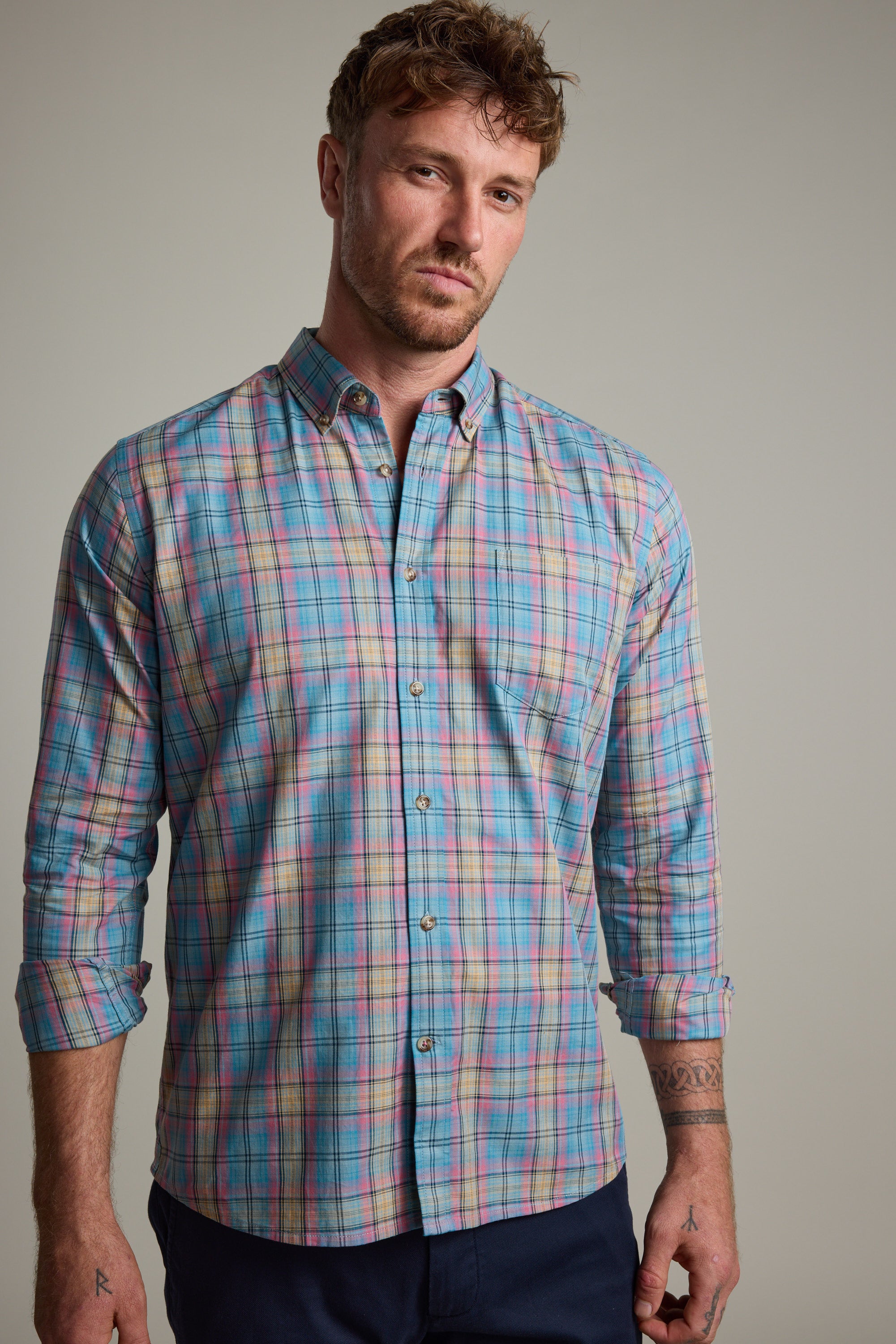 A man with short, curly hair and arm tattoos wears the Barkers Hayne Check Shirt, tailored fit with rolled sleeves, paired with dark pants, standing against a plain light background.
