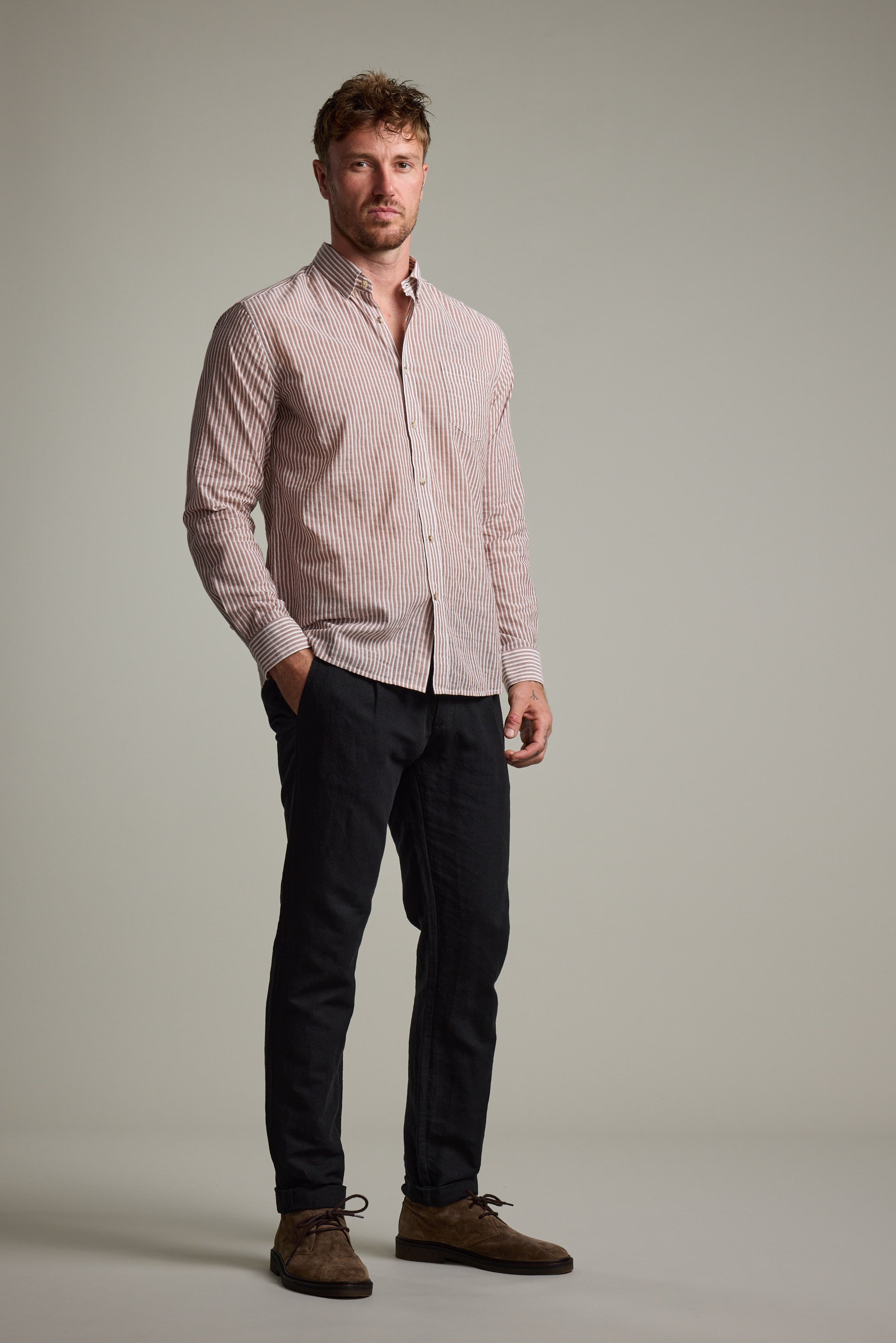 A man stands against a plain background wearing the Barkers Sumter Stripe Shirt—a lightweight, light pink and white striped tailored fit—paired with dark pants and brown shoes, hands in pockets and a neutral expression.