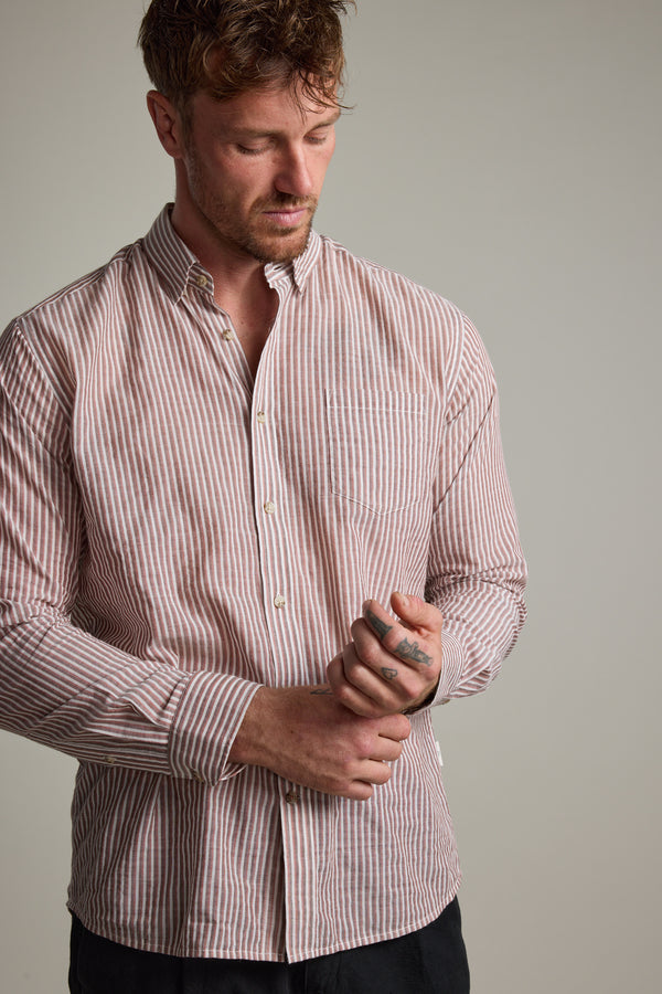 A man in a Barkers Sumter Stripe Shirt, a lightweight red-and-white striped long sleeve button-down, looks down as he adjusts his sleeve against a plain neutral background.