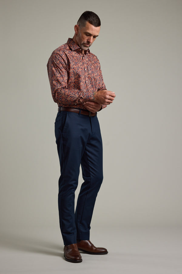 A man, dressed in a Barkers Paisley Business Shirt in classic fit, buttons his cuff against a simple backdrop. He completes the look with navy pants and brown dress shoes for an effortlessly sharp style.