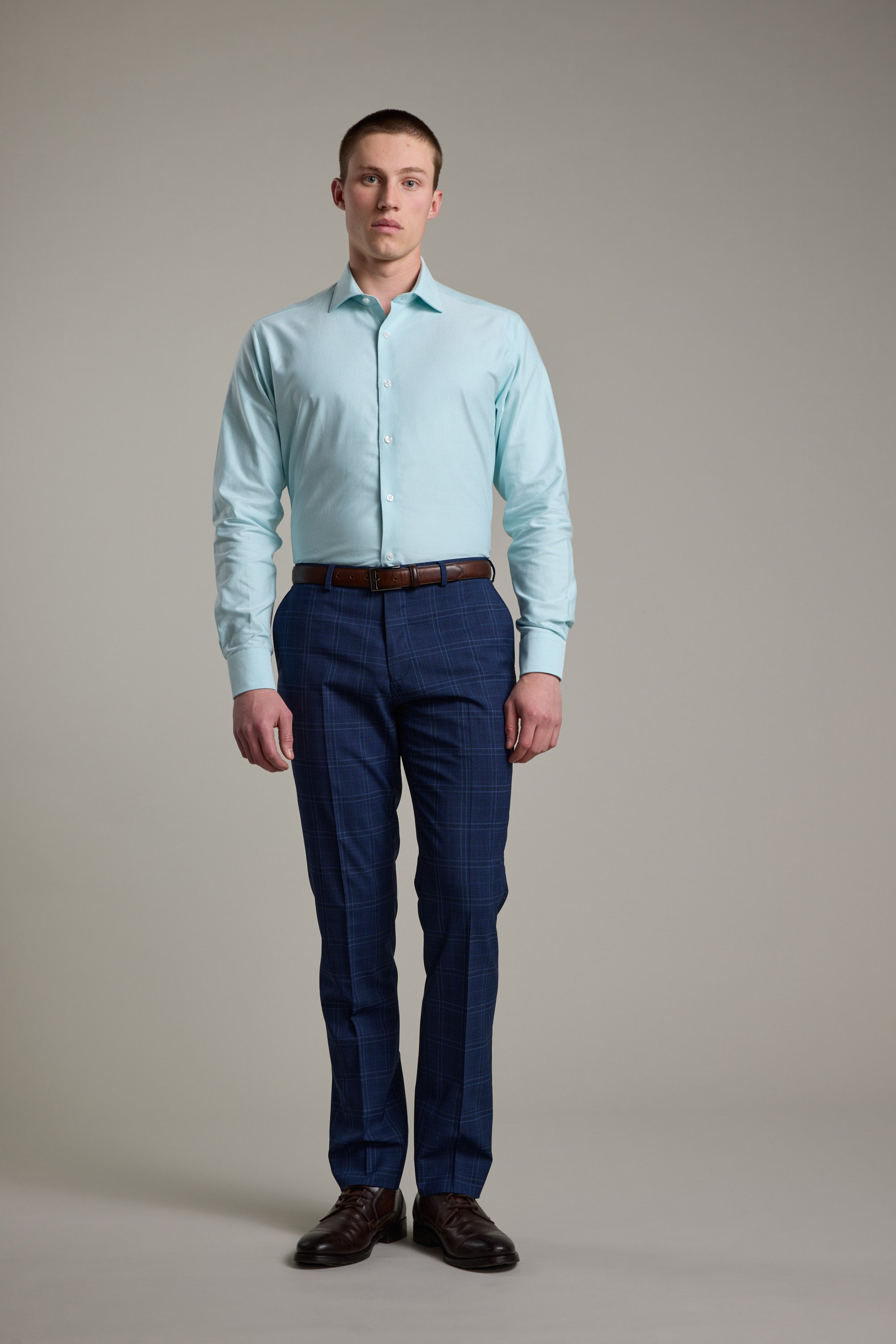 A man stands facing forward against a plain background, wearing the Barkers Edgar Texture Business Shirt in light blue, blue plaid dress pants, a brown belt, and brown dress shoes.