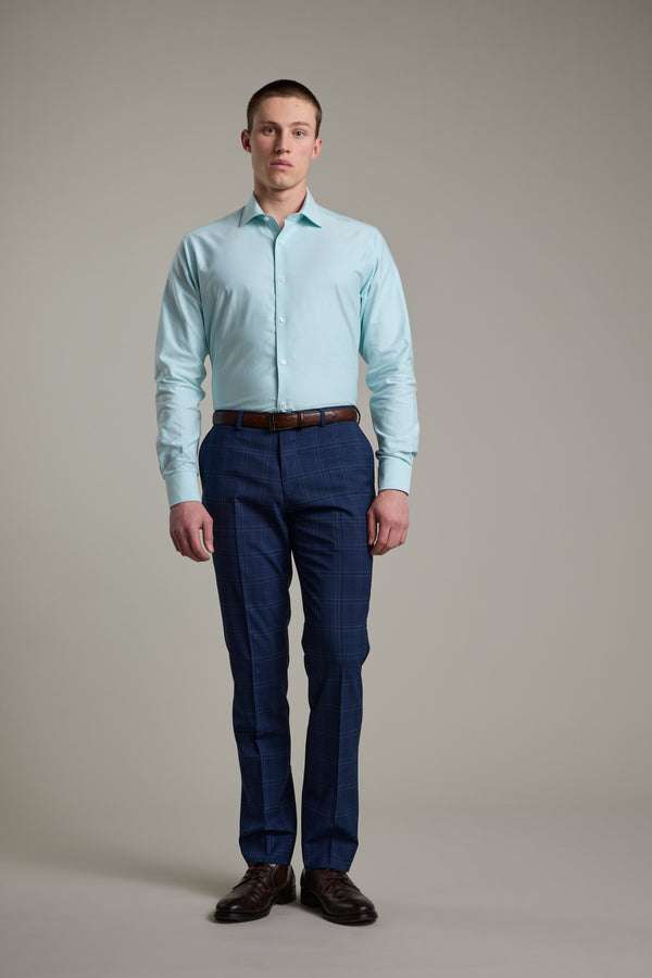 A man stands facing forward against a plain background, wearing the Barkers Edgar Texture Business Shirt in light blue, blue plaid dress pants, a brown belt, and brown dress shoes.