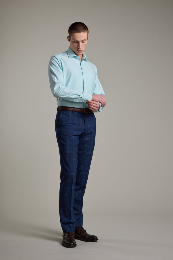 A man stands against a plain background, looking down as he adjusts the cuff of his Barkers Edgar Texture Business Shirt. He pairs it with blue plaid dress pants, a brown belt, and dark brown shoes.